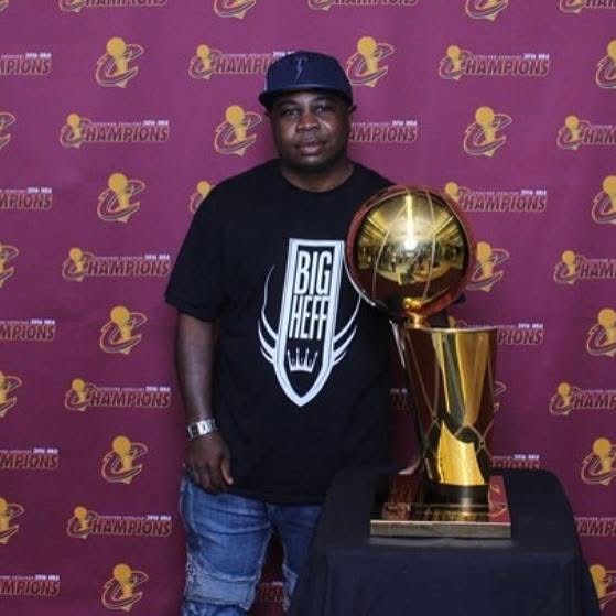 Man standing next to a large golden trophy, wearing a black T-shirt and cap, in front of a maroon background with logos.