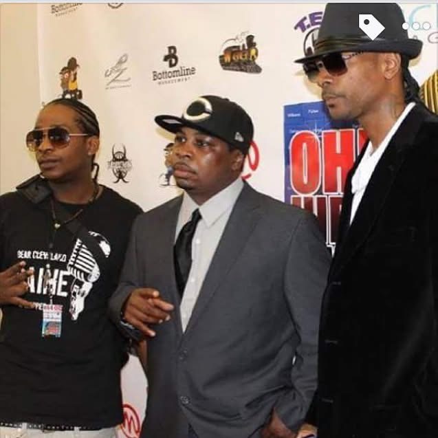 Three men in stylish clothing and sunglasses standing together at an event with a backdrop of logos and posters.