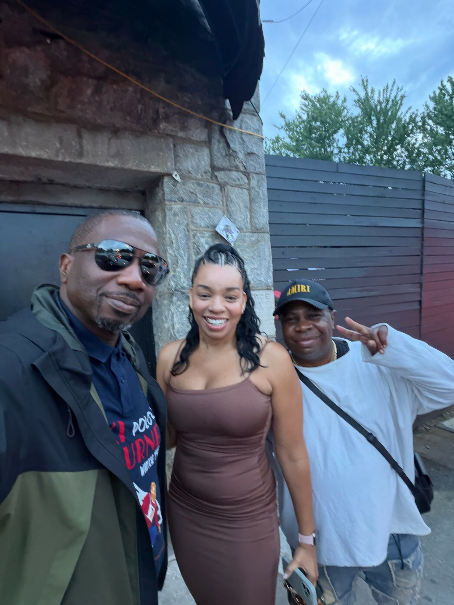 Three people smiling outdoors, standing close together near a stone wall and fence, with trees and sky in background.