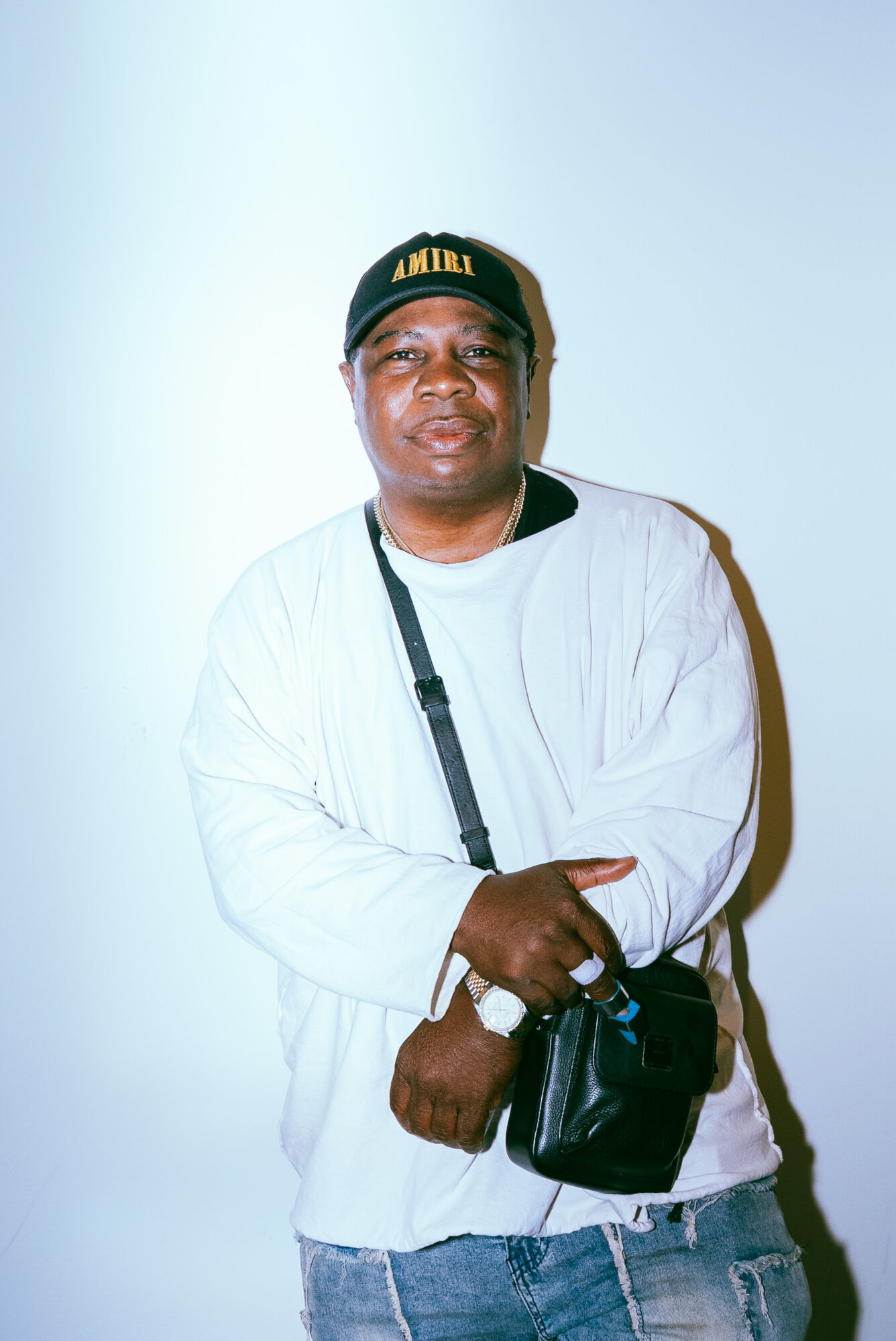 Man wearing a black cap, white jacket, and jeans, holding a black bag, standing against a light background.