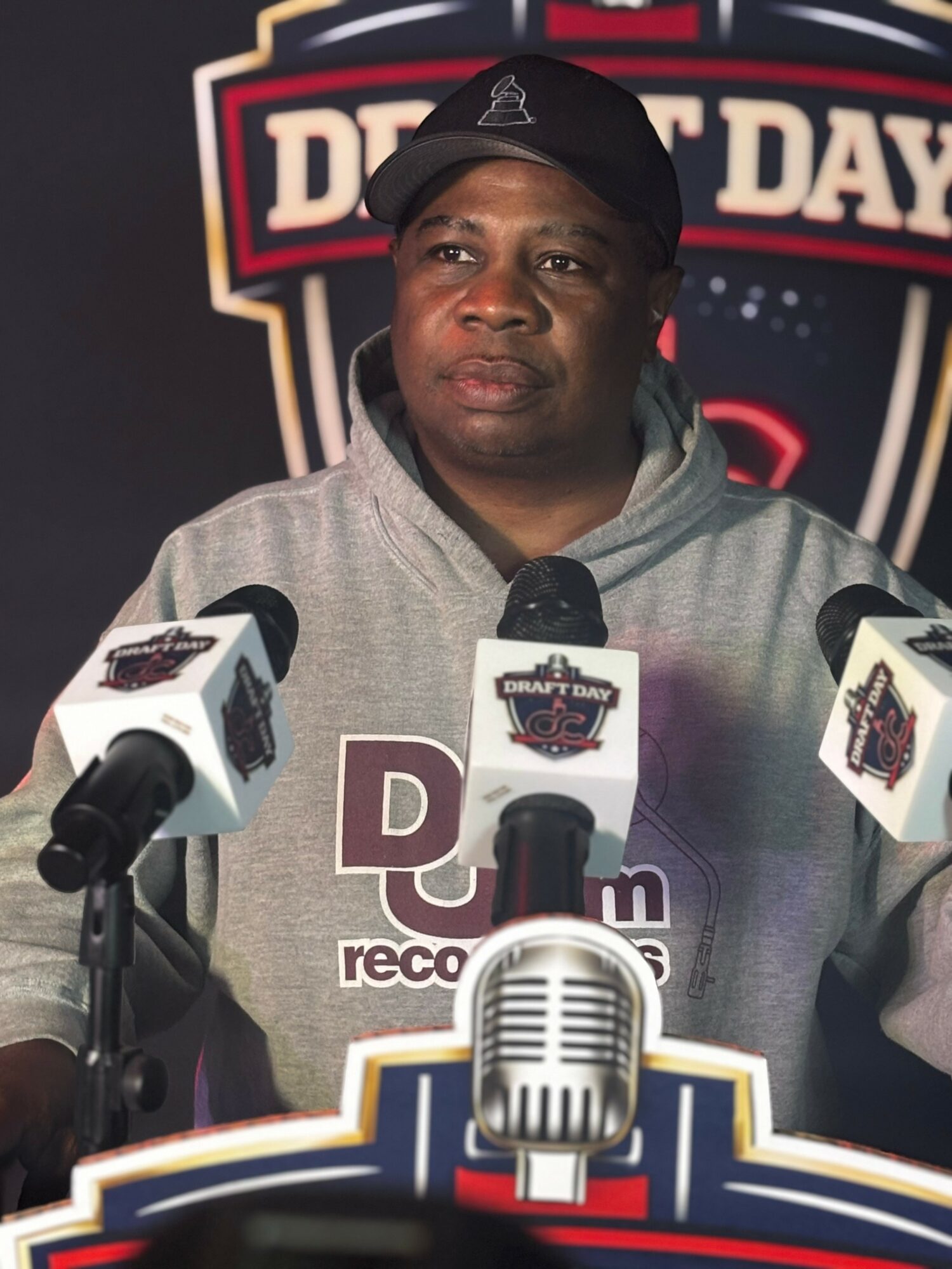 Man in gray hoodie and black cap speaking at a press conference with microphones, background features a logo, neutral expression.