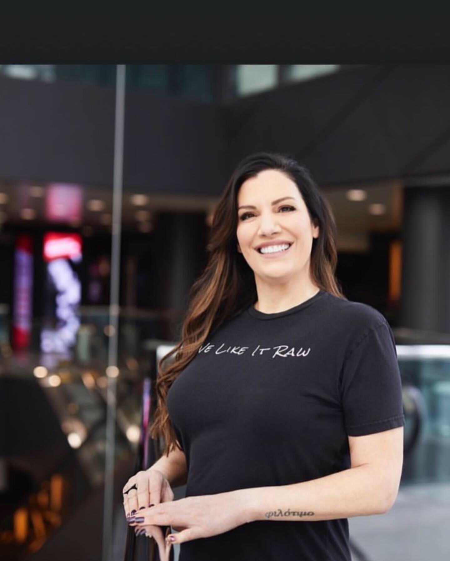 Smiling woman with long dark hair in a black T-shirt standing indoors, background with glass and lights.