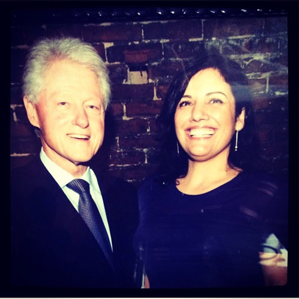 Two people smiling in front of a brick wall, one older man and one woman with dark hair, both dressed formally.