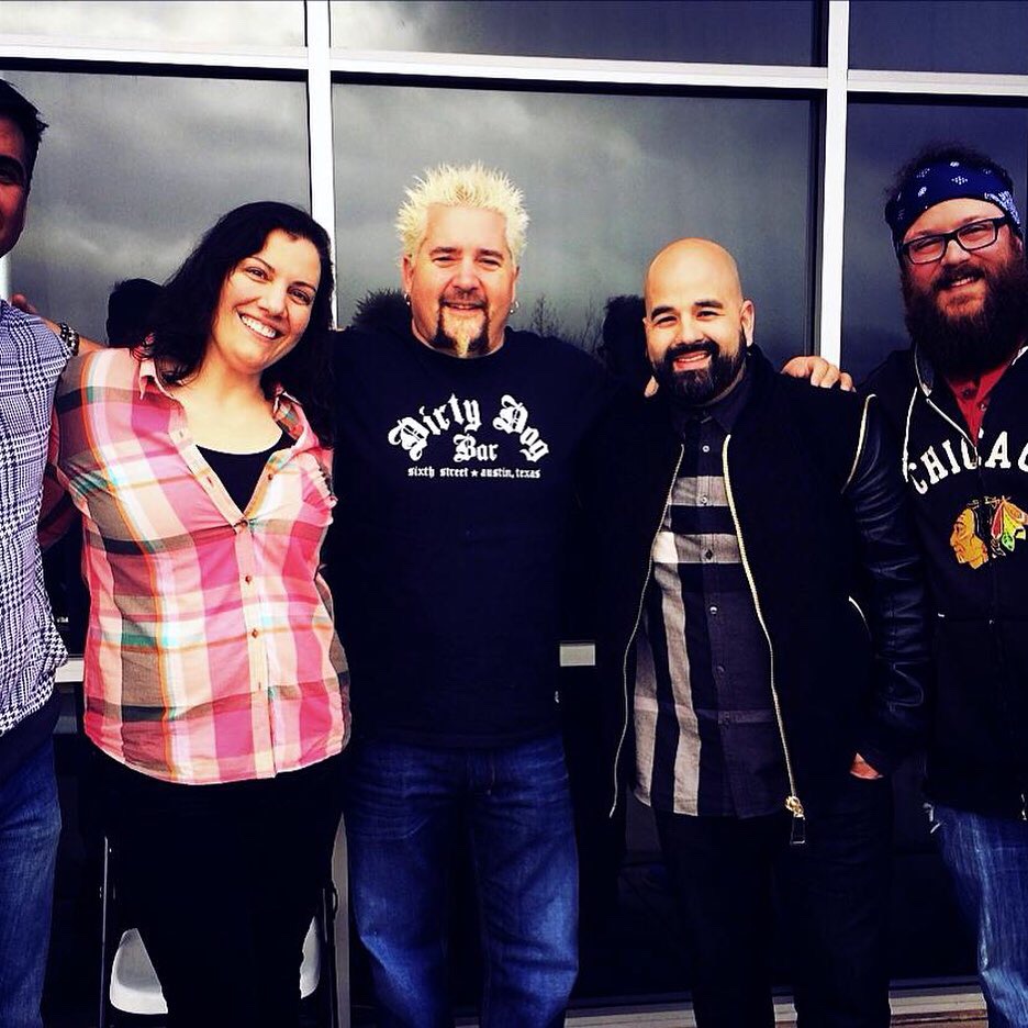 Group of five people smiling outdoors in front of a glass building, diverse in appearance, casual clothing, some with beards and glasses.