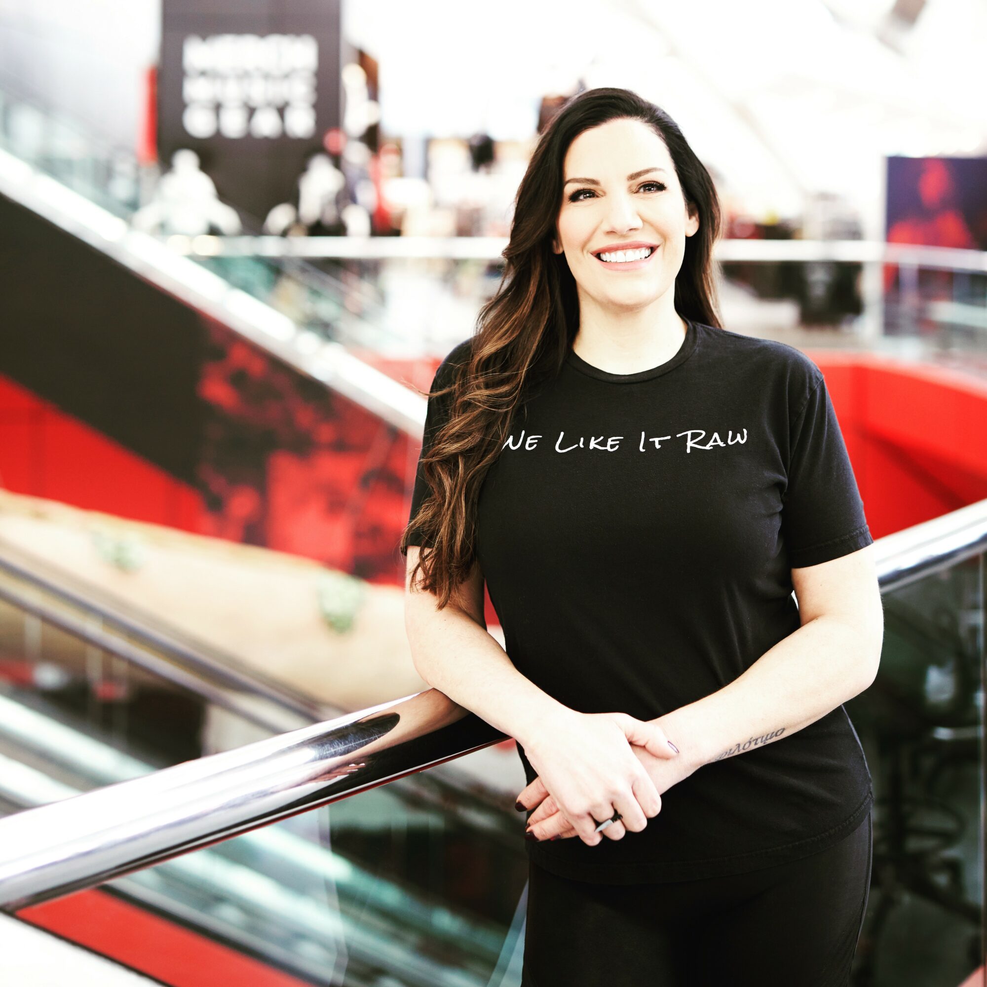 Smiling woman with long dark hair standing indoors, wearing a black T-shirt with white text, leaning on a railing.