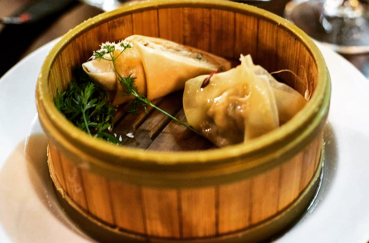 Two dumplings with herbs in a round wooden steamer basket on a white plate.