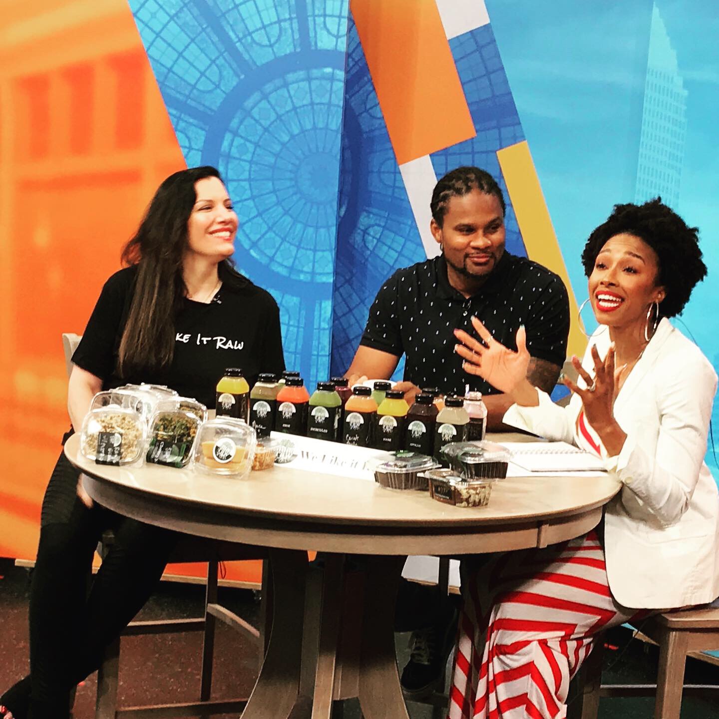 Three people sit around a table with jars of food, smiling and talking, in a colorful studio setting.