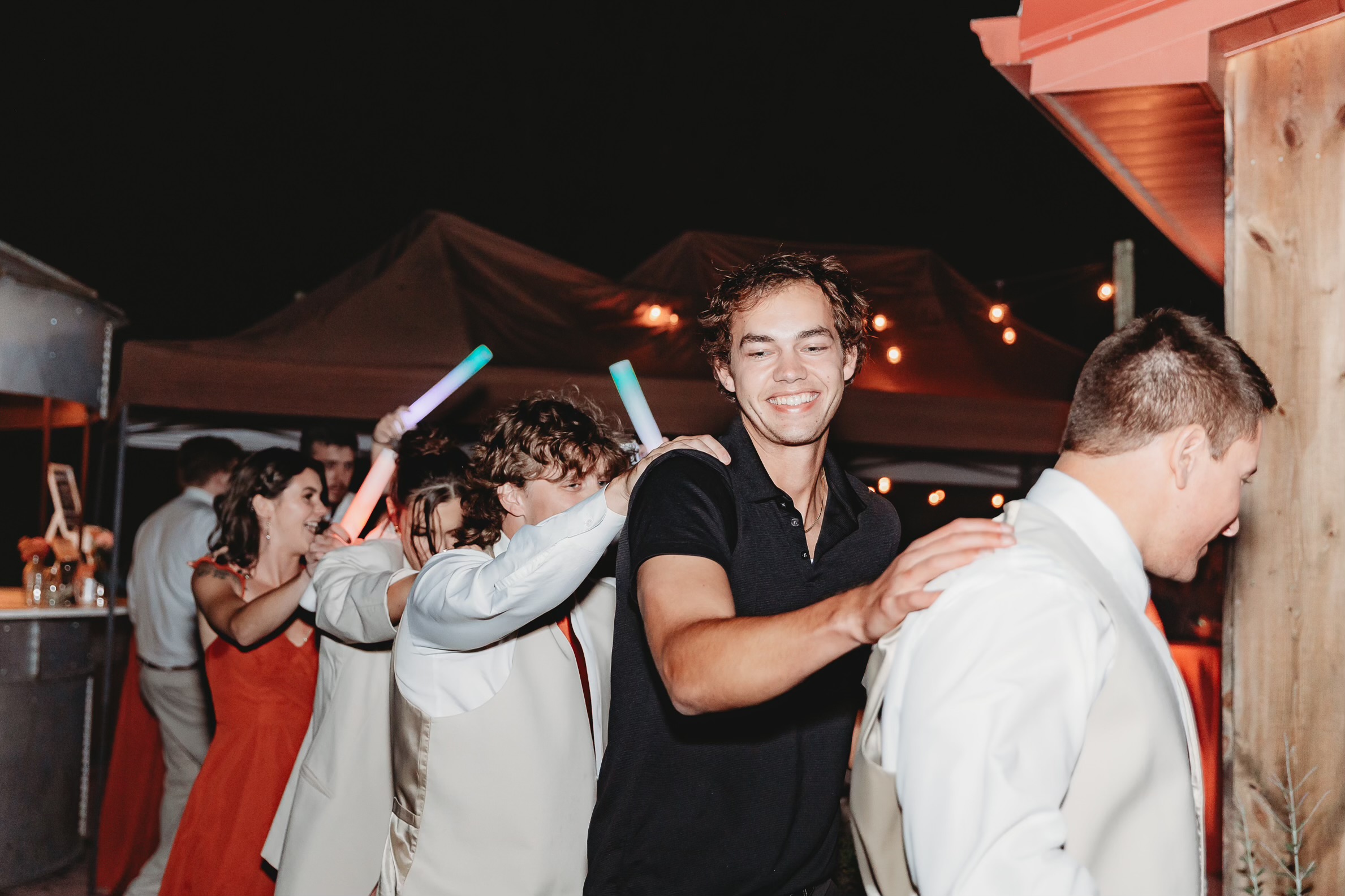 Group of people celebrating outdoors at night, smiling, with some holding glow sticks, under tents and string lights.