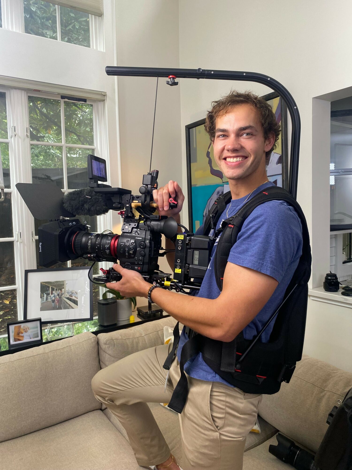 Young man with camera equipment sitting on a beige couch in a room with large windows and a door, smiling at the camera.