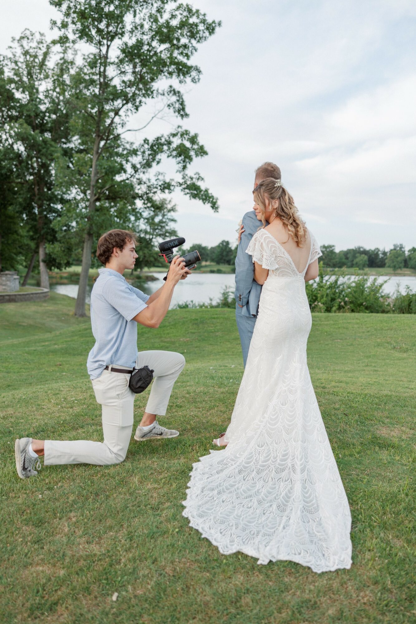 A person kneels on grass taking a photo of a woman in a white wedding dress outdoors near a lake, with trees and sky in background.