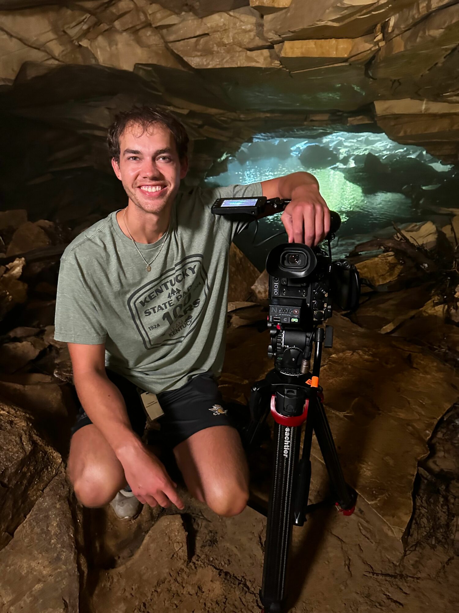 Person sitting in a rocky cave holding a camera on a tripod, smiling, with water visible in the background.
