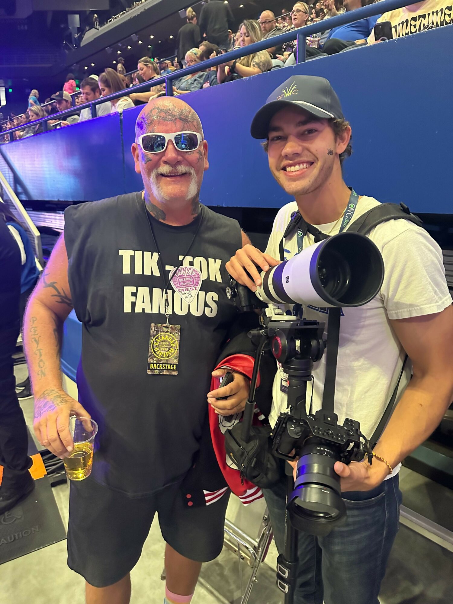 Two men posing with a camera and a drink, one wearing sunglasses and a T-shirt, the other wearing a cap and holding a camera, in an indoor setting with a crowd in the background.