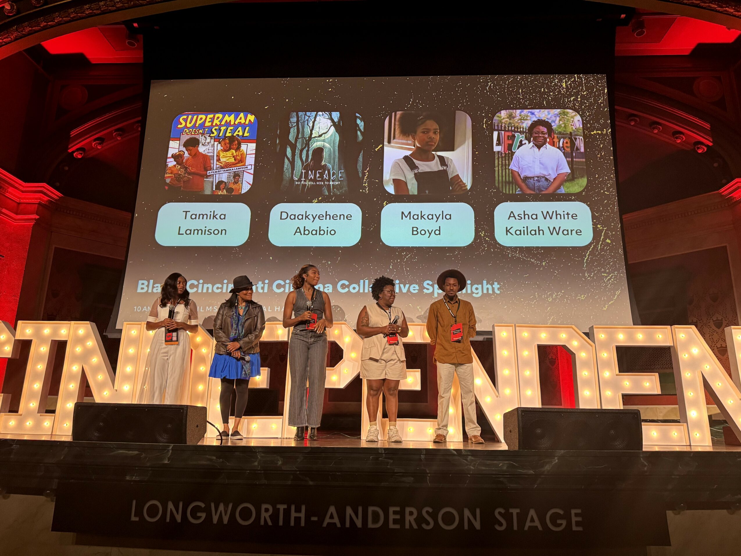 Six people stand on stage in front of a large screen displaying children's photos and names, with illuminated letters spelling 'TALENT' behind them.
