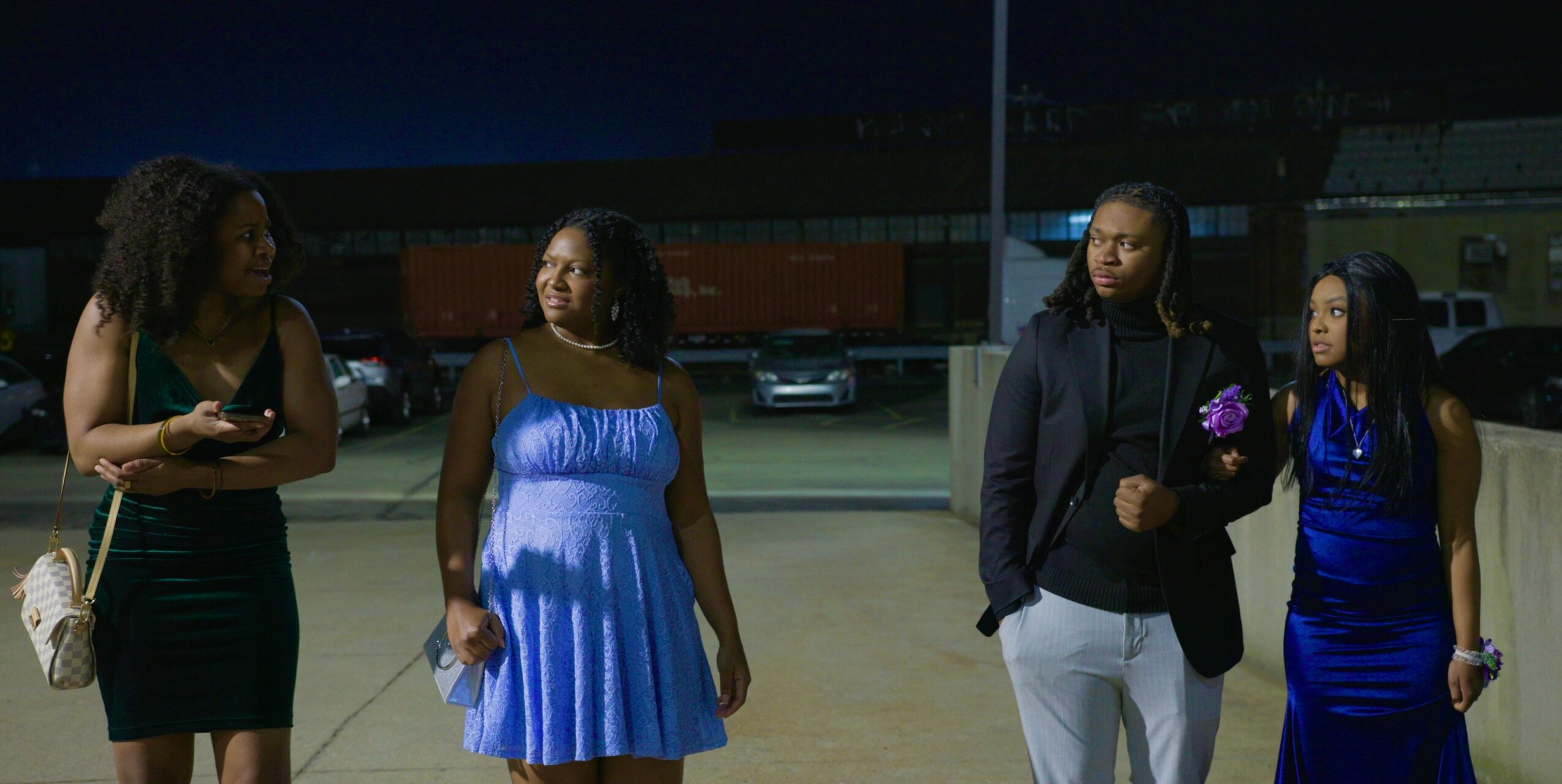 Four people standing outdoors at night, dressed formally, with a parking lot and stadium in background.