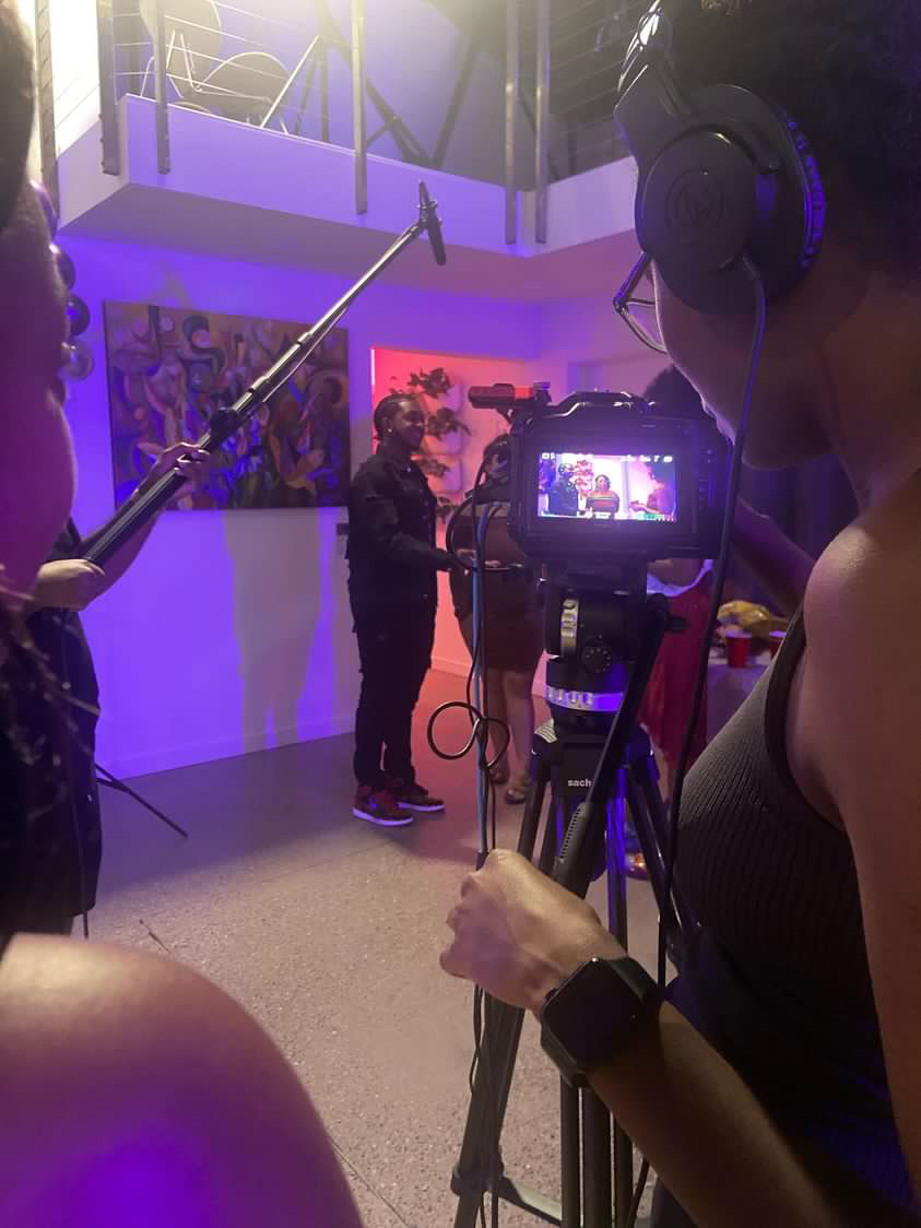 Filming a person speaking in front of a colorful painting, with crew members and equipment visible, in a studio setting.