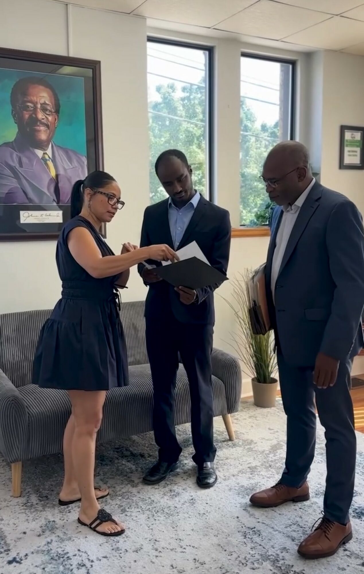 Three people standing indoors, two men in suits and a woman in a dress, looking at a document together.
