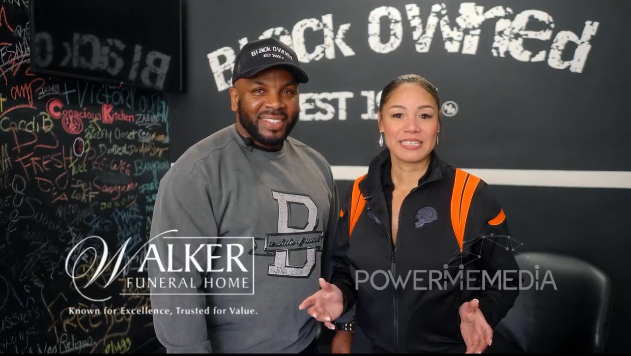Two people shaking hands in front of a black wall with white text, one wearing a gray sweatshirt and the other a black jacket with orange straps.