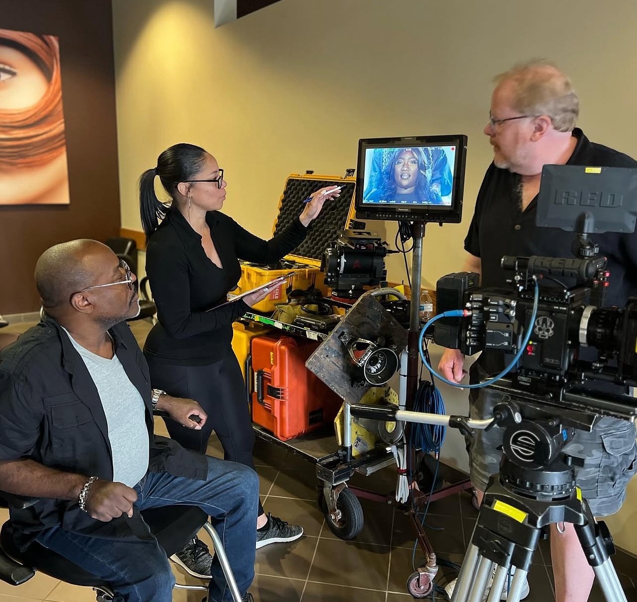 Three people in a filming studio with cameras and monitors, one woman explaining to a man, another man seated nearby.