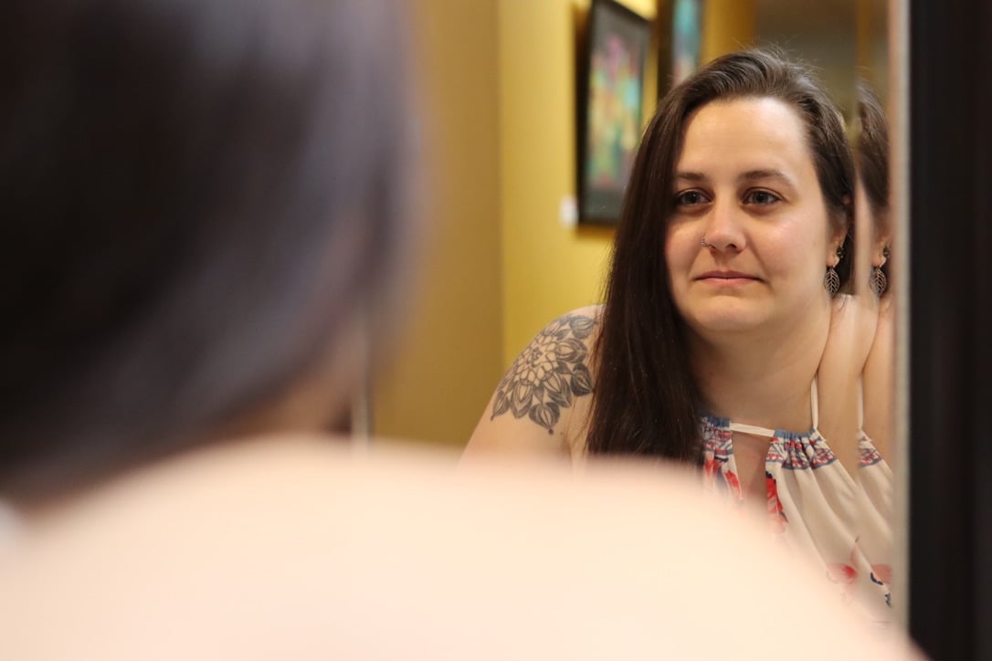 Woman looking at her reflection in a mirror, smiling slightly, with a yellow wall and framed pictures in the background.