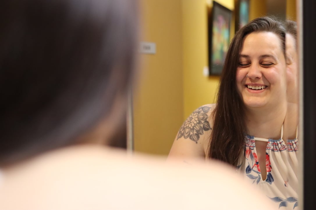 Smiling woman with long dark hair and a tattoo on her shoulder, wearing a patterned top, in a room with yellow walls and framed pictures.