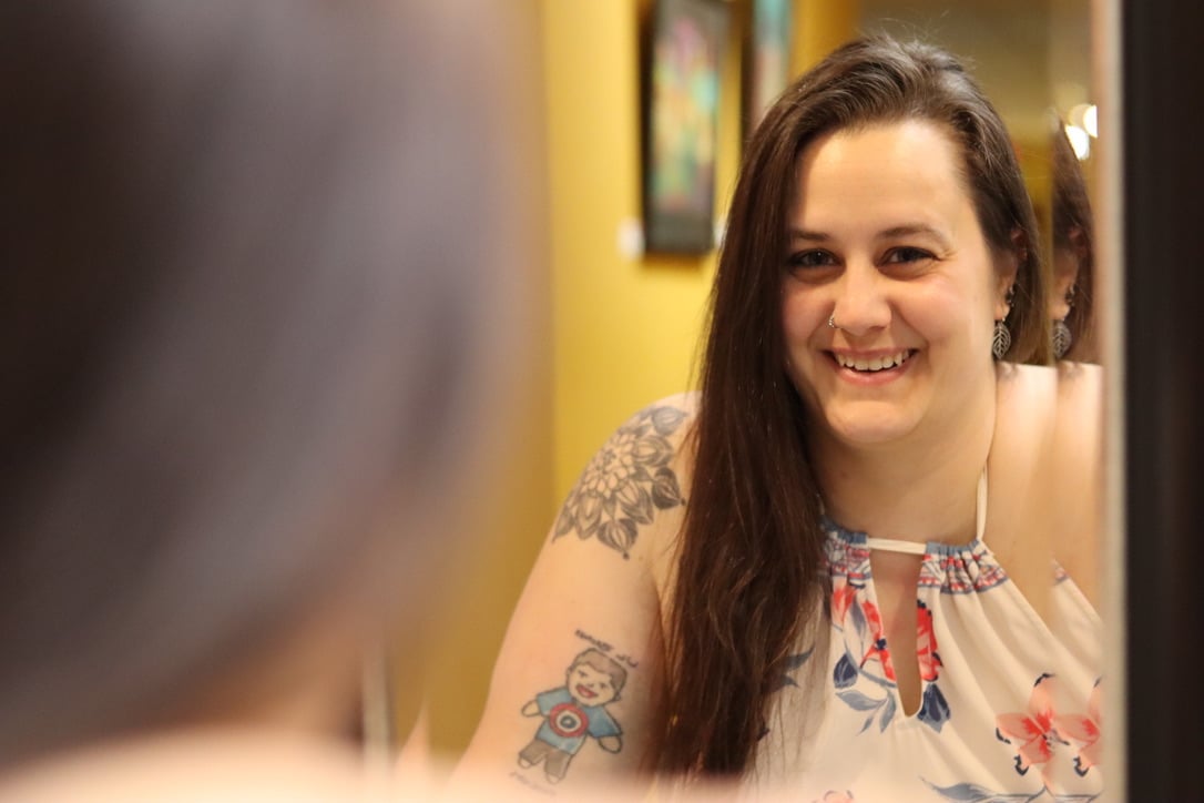 Smiling woman with long dark hair and a tattoo on her arm, wearing a floral top, in a brightly colored room.