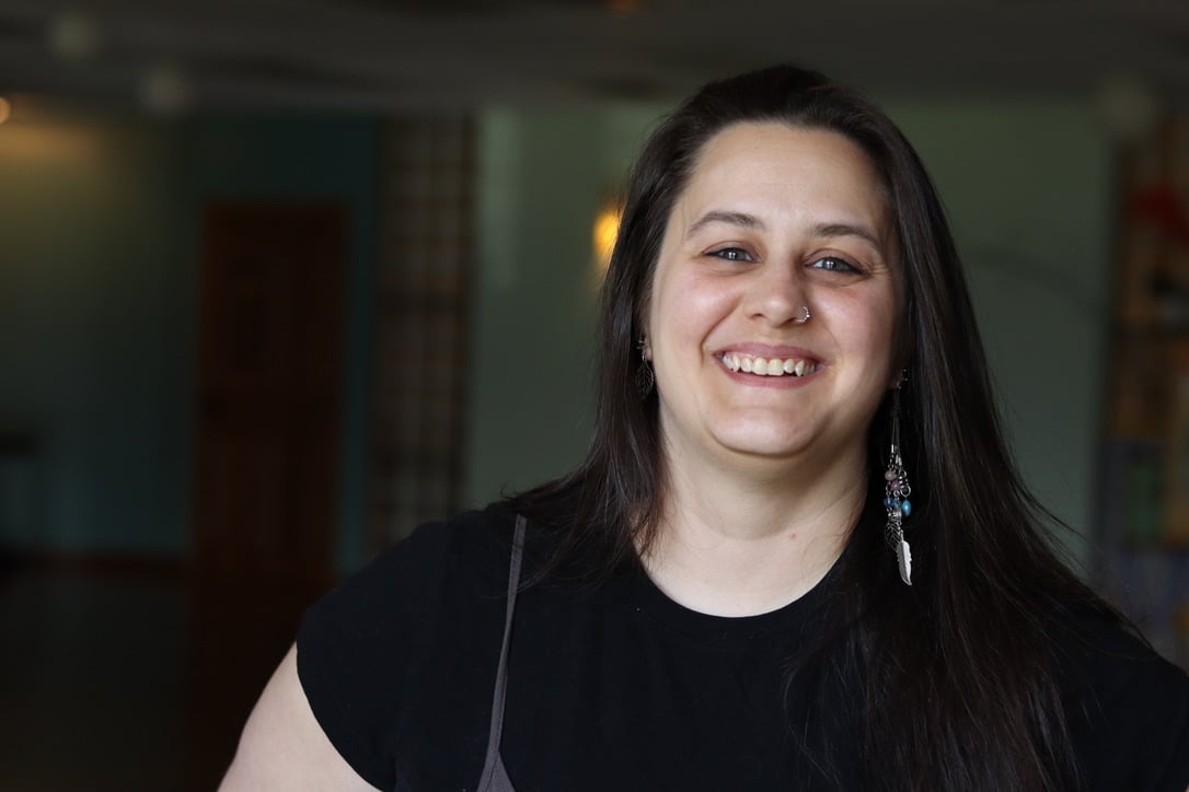 Smiling woman with long dark hair wearing a black shirt, indoors with blurred background.