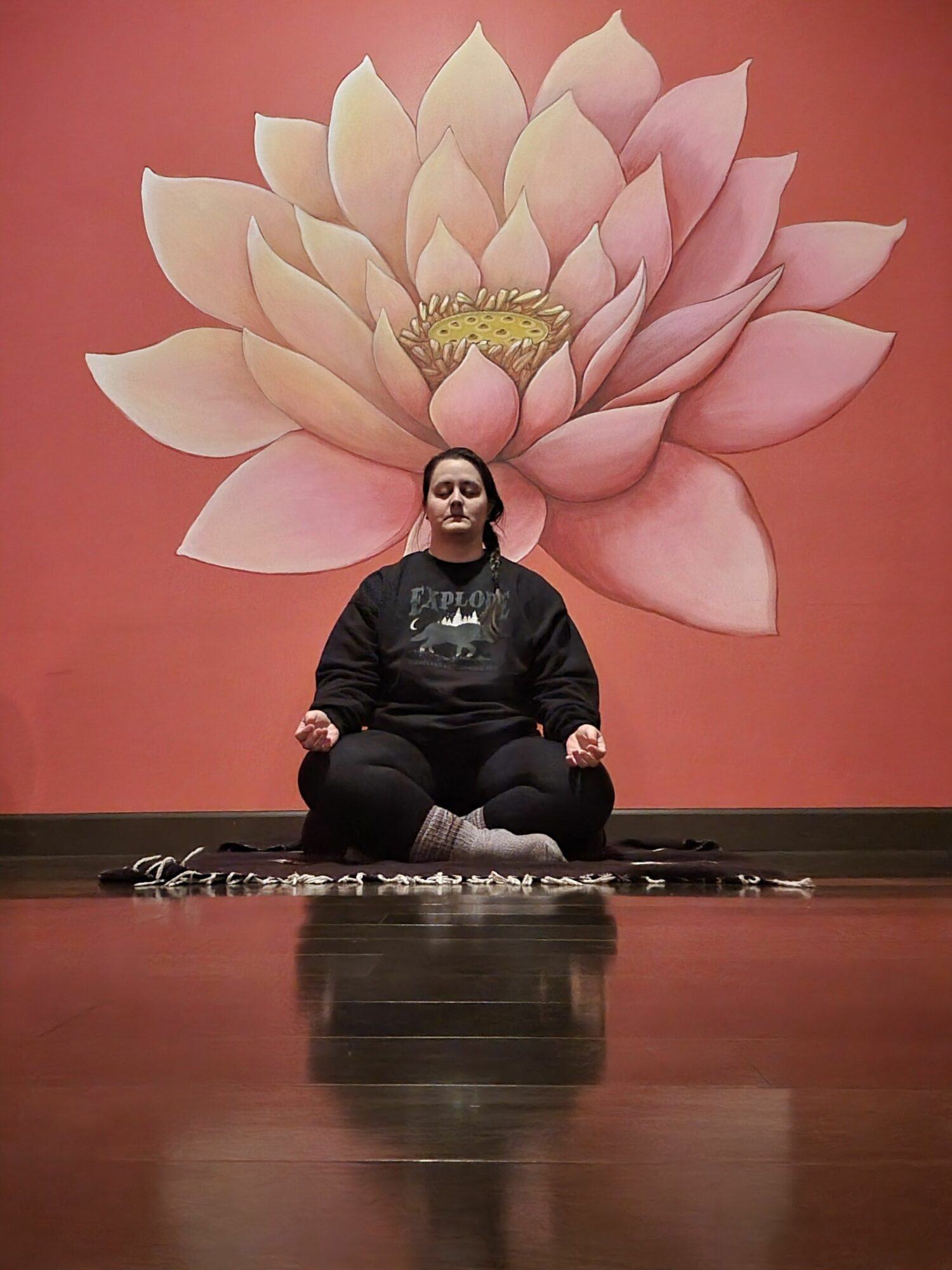 Person sitting at a table with a large pink lotus flower mural behind them, facing forward, with a neutral expression.