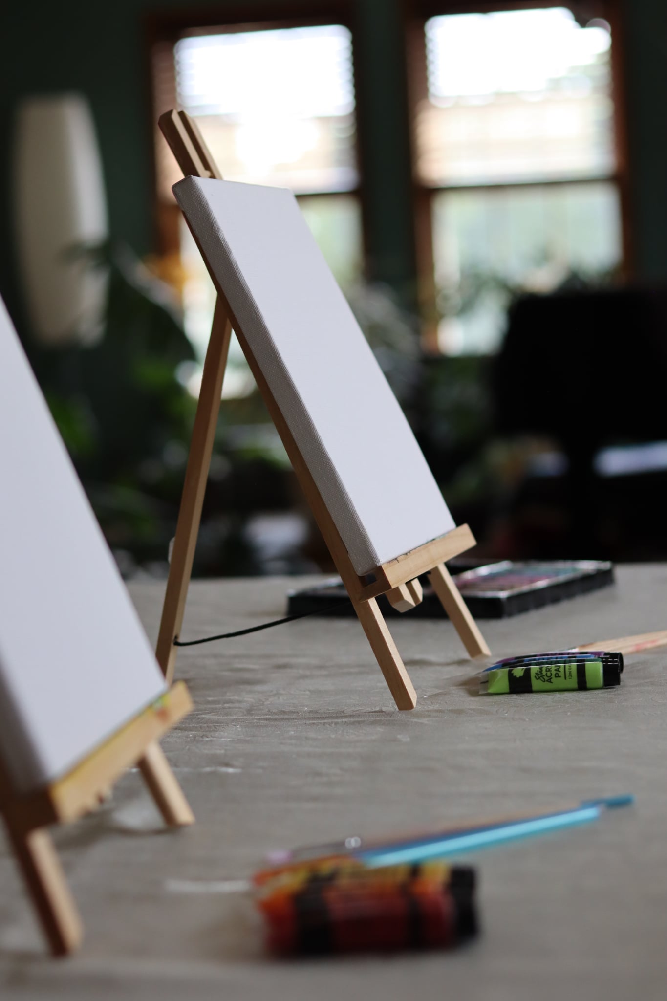 Small easel with blank canvas on a table, with a green marker and colorful supplies nearby, in a room with windows.