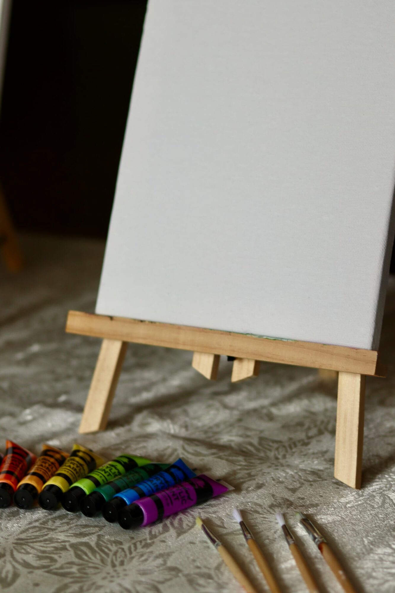 Blank white canvas on small wooden easel with colored chalks and pens on textured surface.