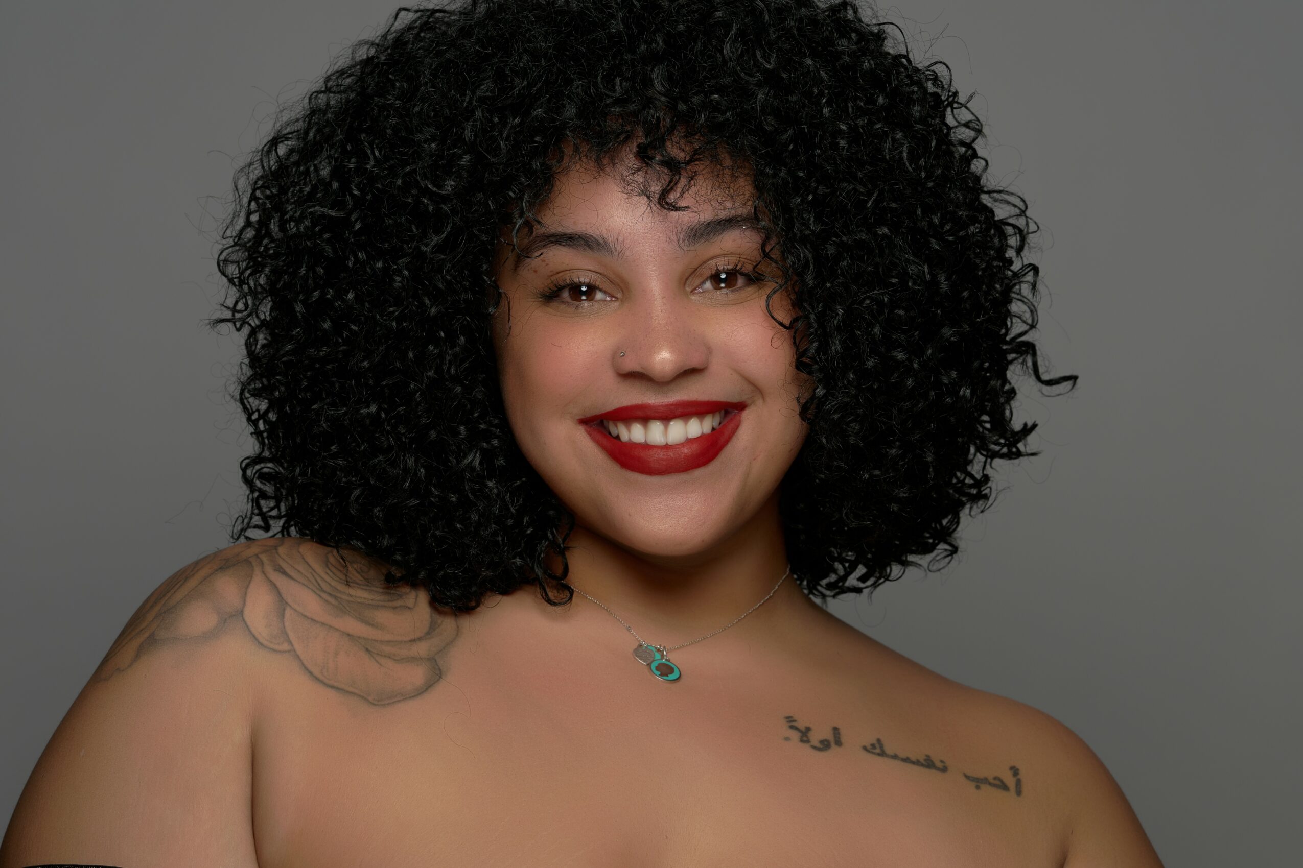 Young woman with curly hair smiling, wearing red lipstick, and a necklace, against a gray background.