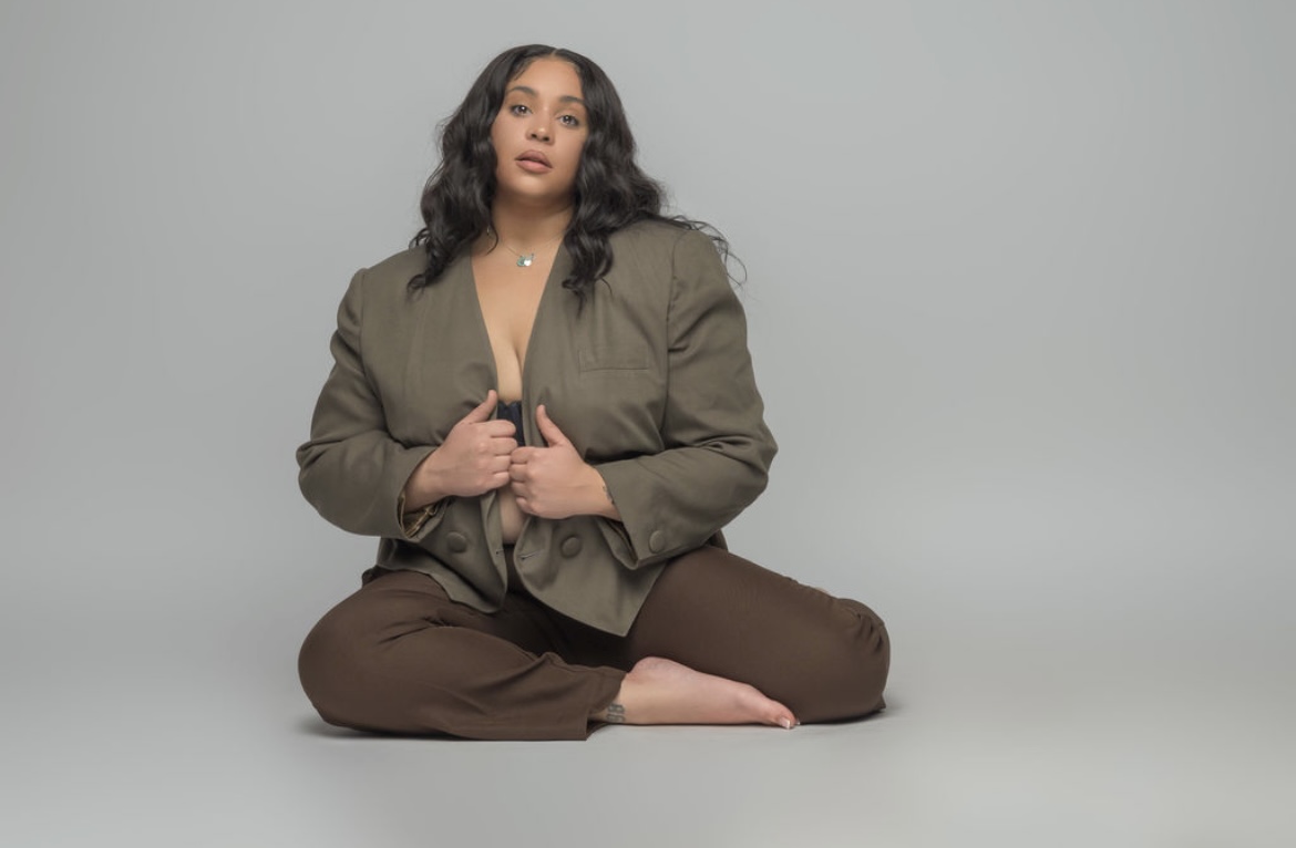 Woman sitting on the floor with legs crossed, wearing an oversized jacket and brown pants, against a plain background.