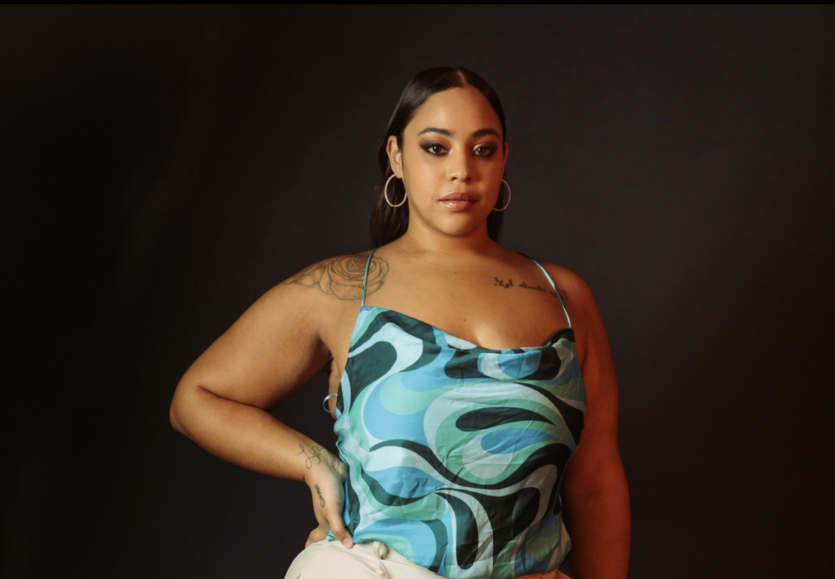 Woman with dark hair and hoop earrings wearing a blue patterned top against a dark background.