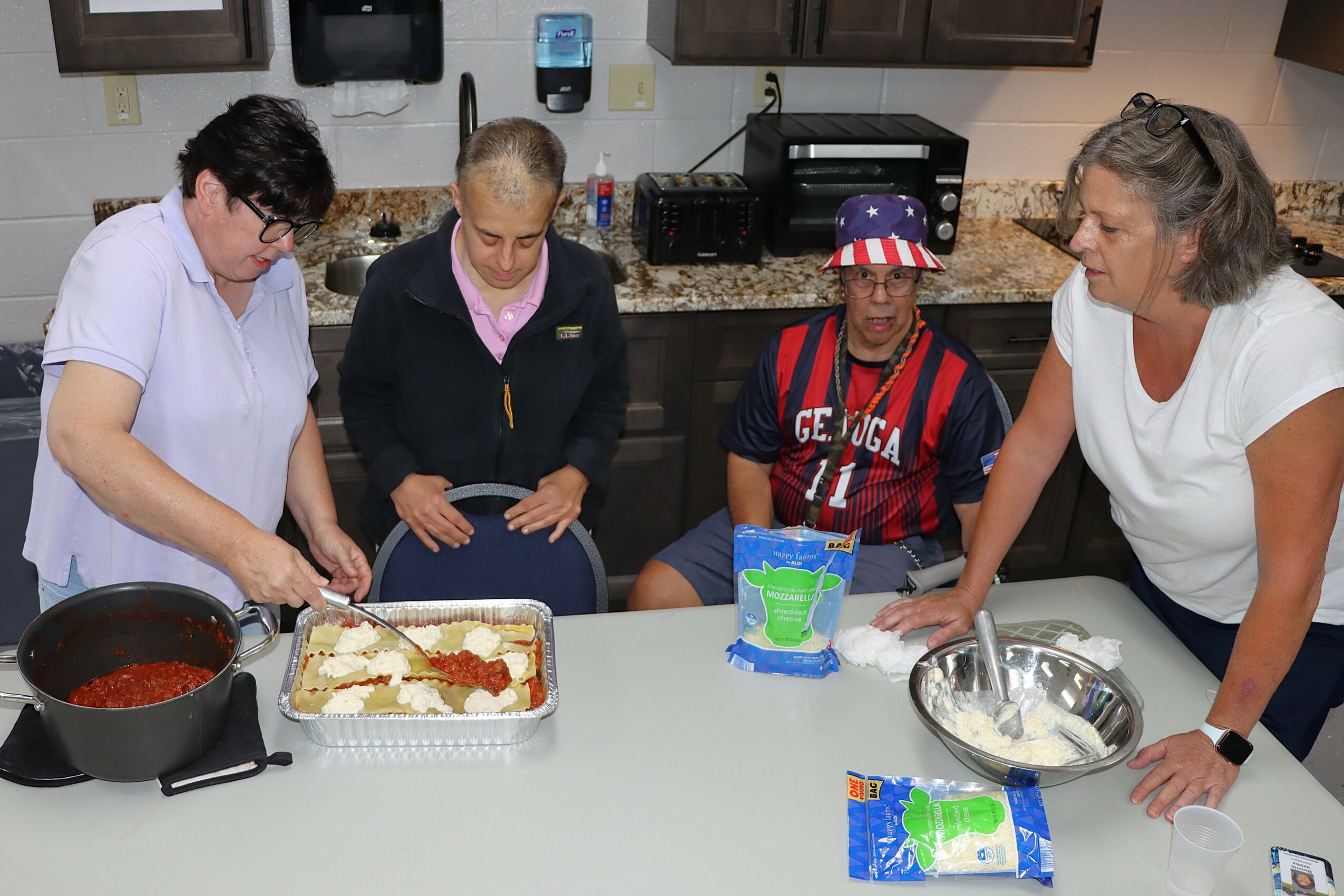 Four women gather around a kitchen counter, preparing food with a baking tray and ingredients, in a home kitchen.