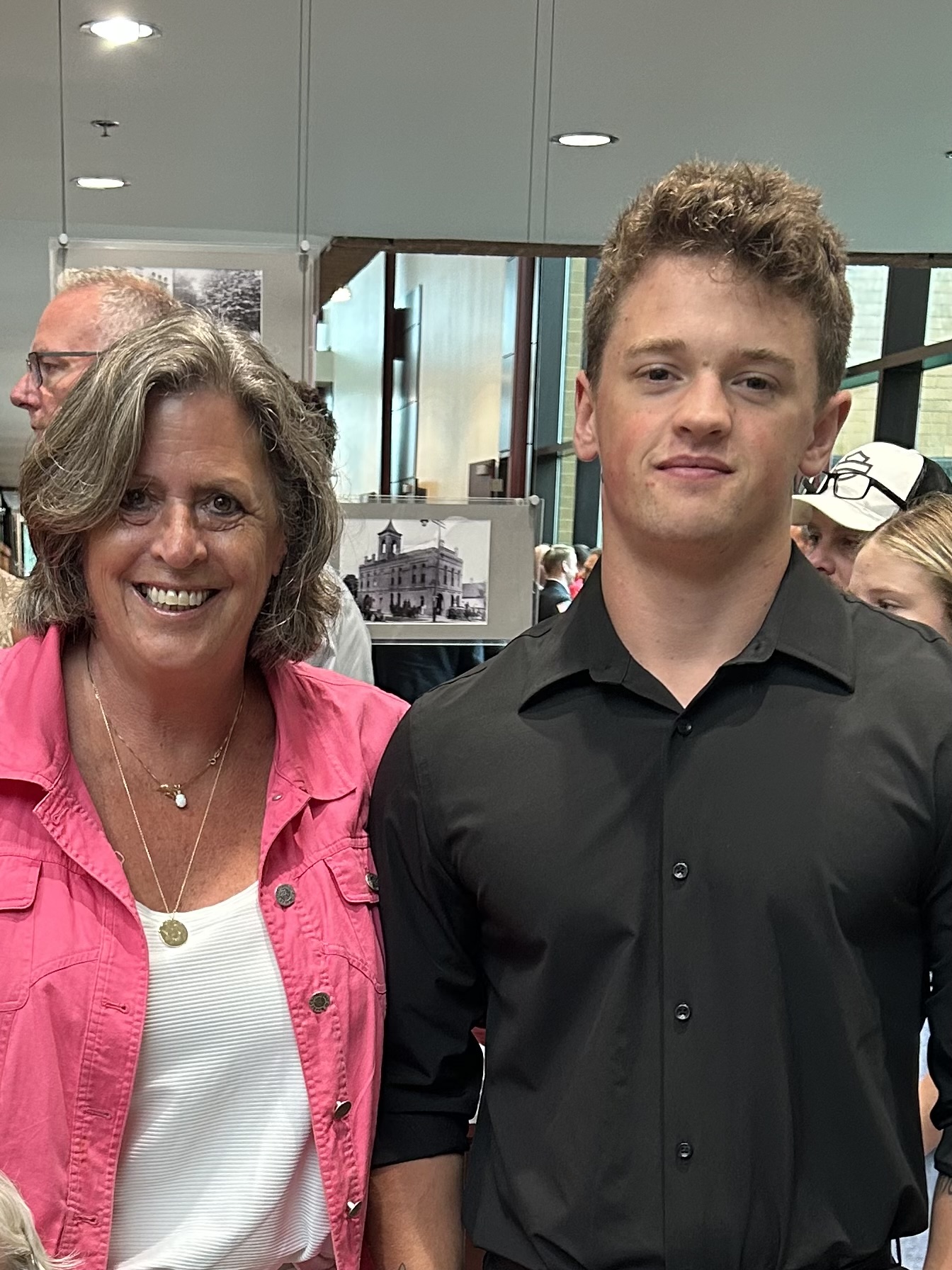 A woman and a young man standing indoors, smiling at the camera, with bookshelves and people in the background.