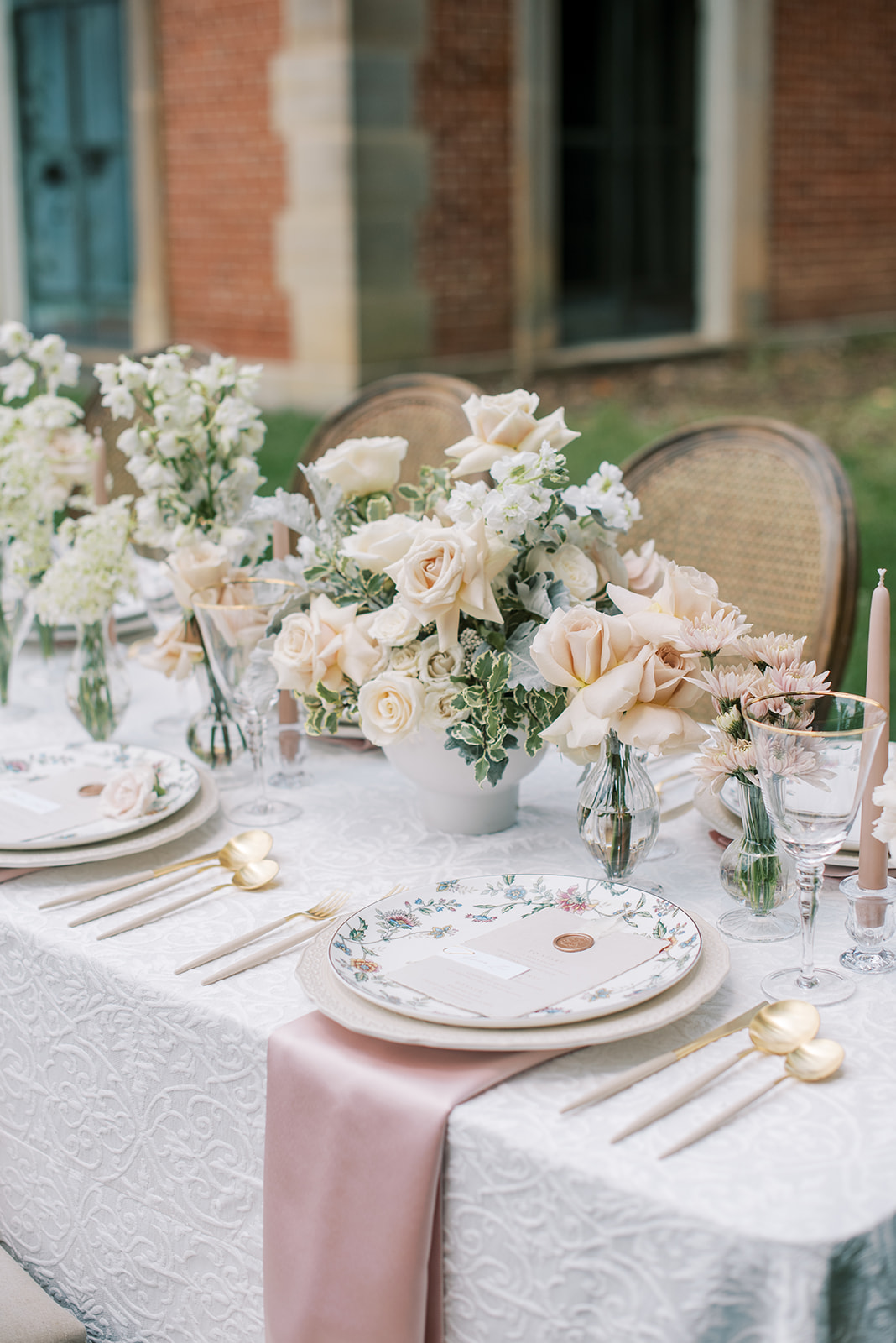 Elegant table setting with floral centerpiece, plates, cutlery, and candles outdoors, brick building in background.