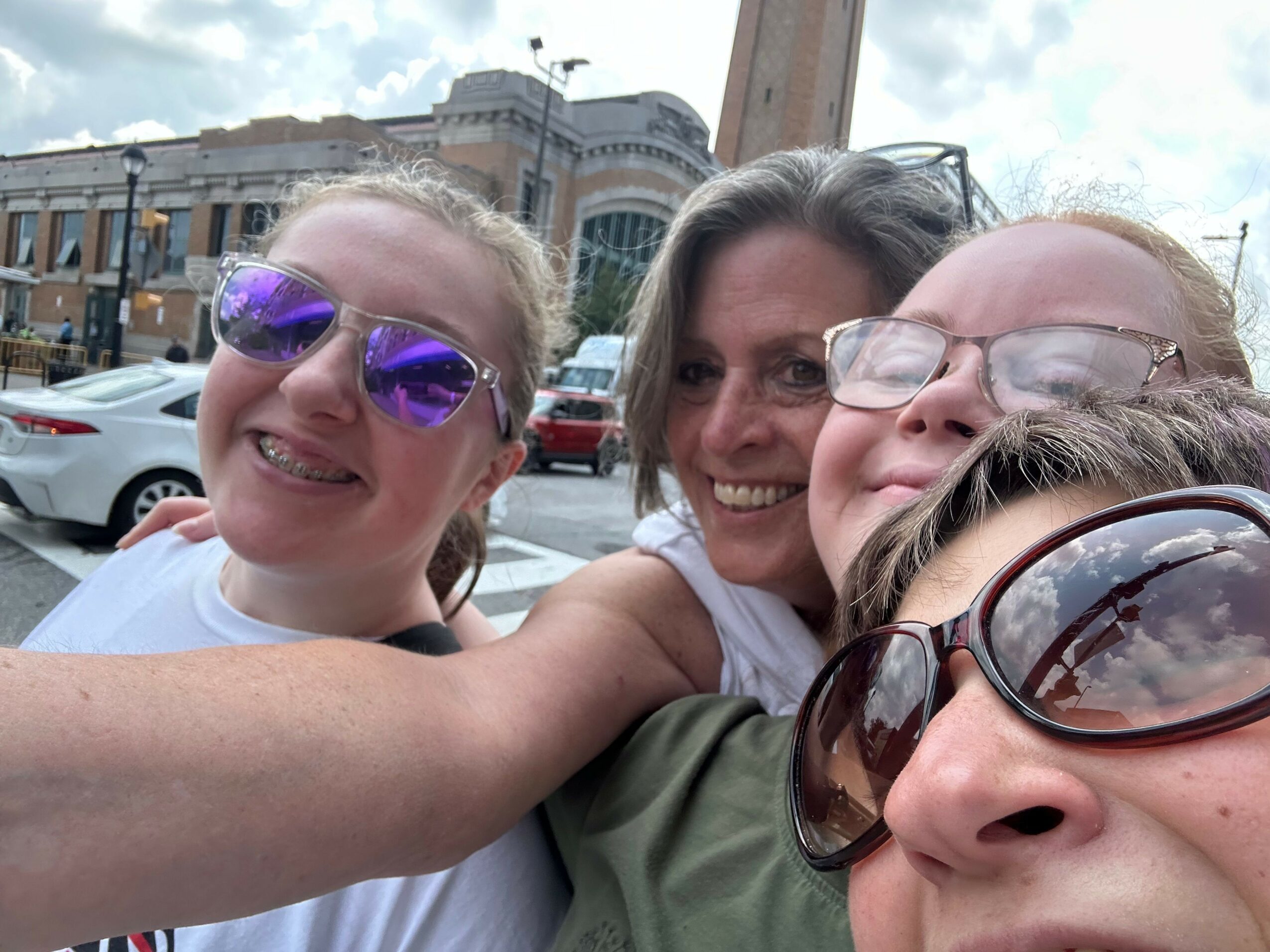 Four people taking a selfie outdoors, smiling, with buildings and cars in the background.
