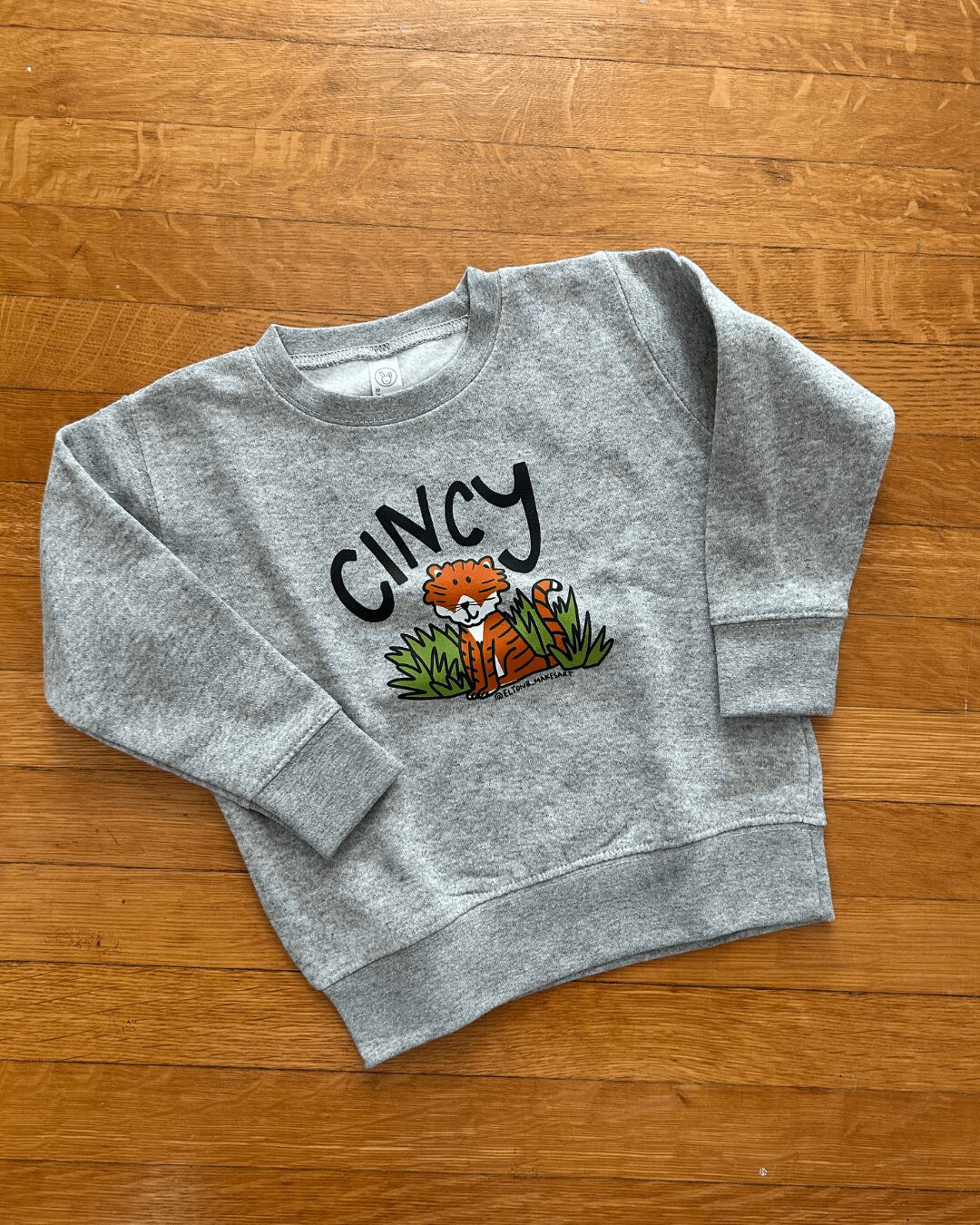 Gray sweatshirt with a tiger and grass design, and the word 'CINCY' above, laid on a wooden surface.