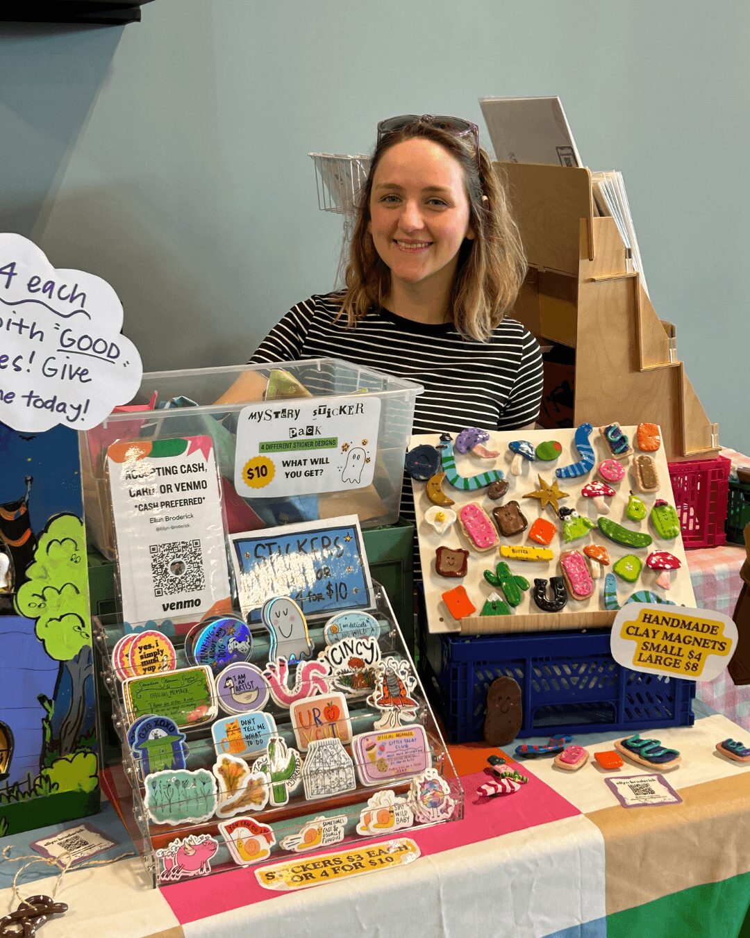 Young woman with long hair behind a table displaying colorful toys and craft items, smiling at the camera.