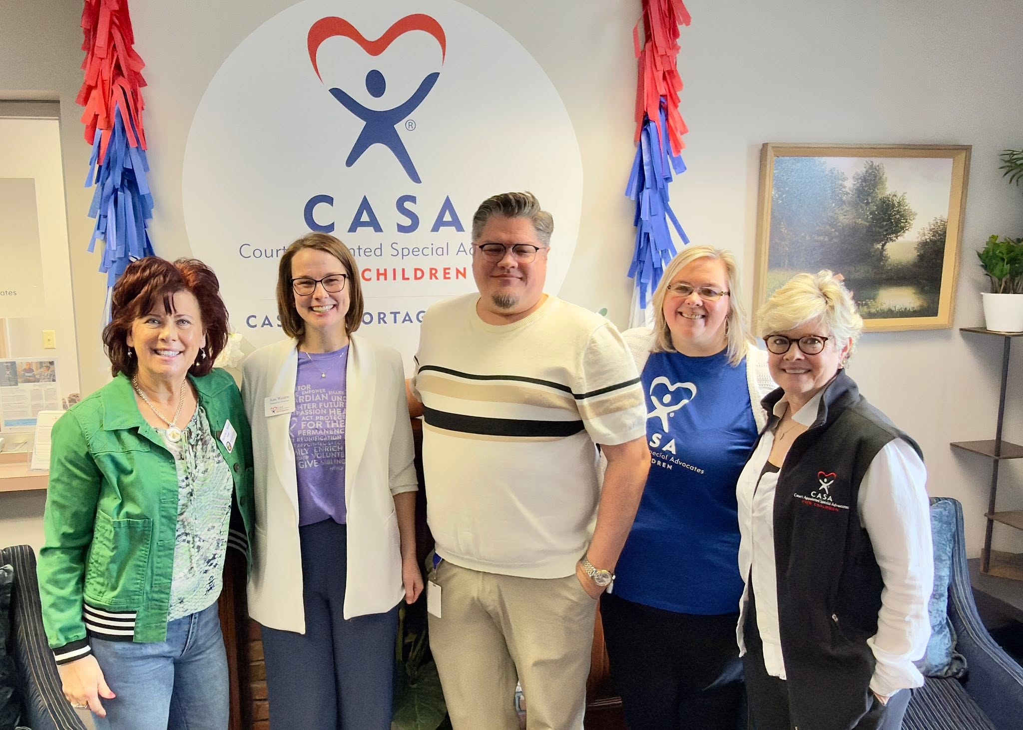 Group of five people standing indoors in front of a CASA banner with red and blue decorations, smiling.