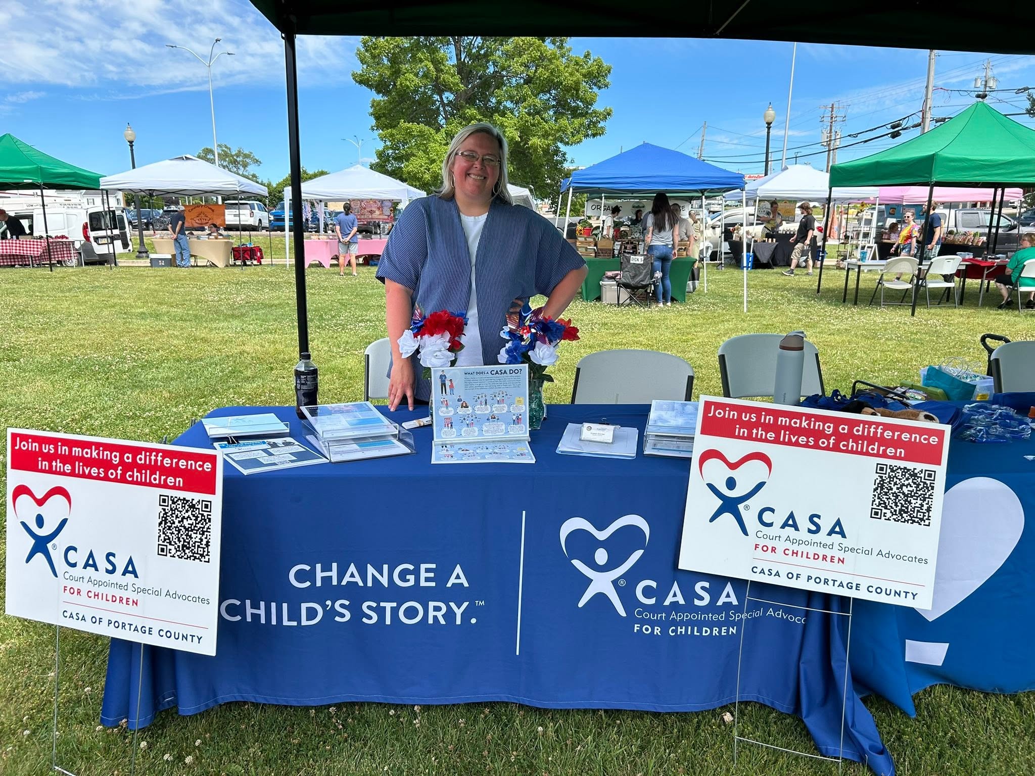 Woman standing behind a table with CASA banners at outdoor event, tents, and people in background.