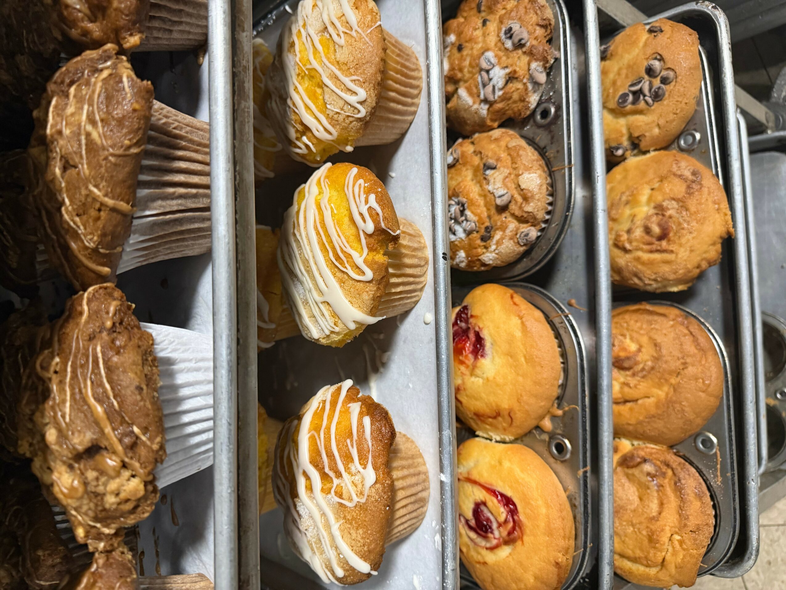 Assorted muffins and pastries on metal racks, some with icing and fruit toppings, in a bakery display.