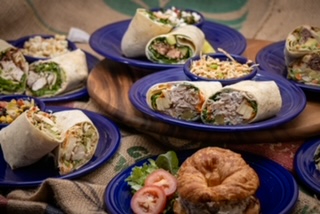 Several wraps on blue plates, a croissant, and sliced tomato on a table with a cloth.