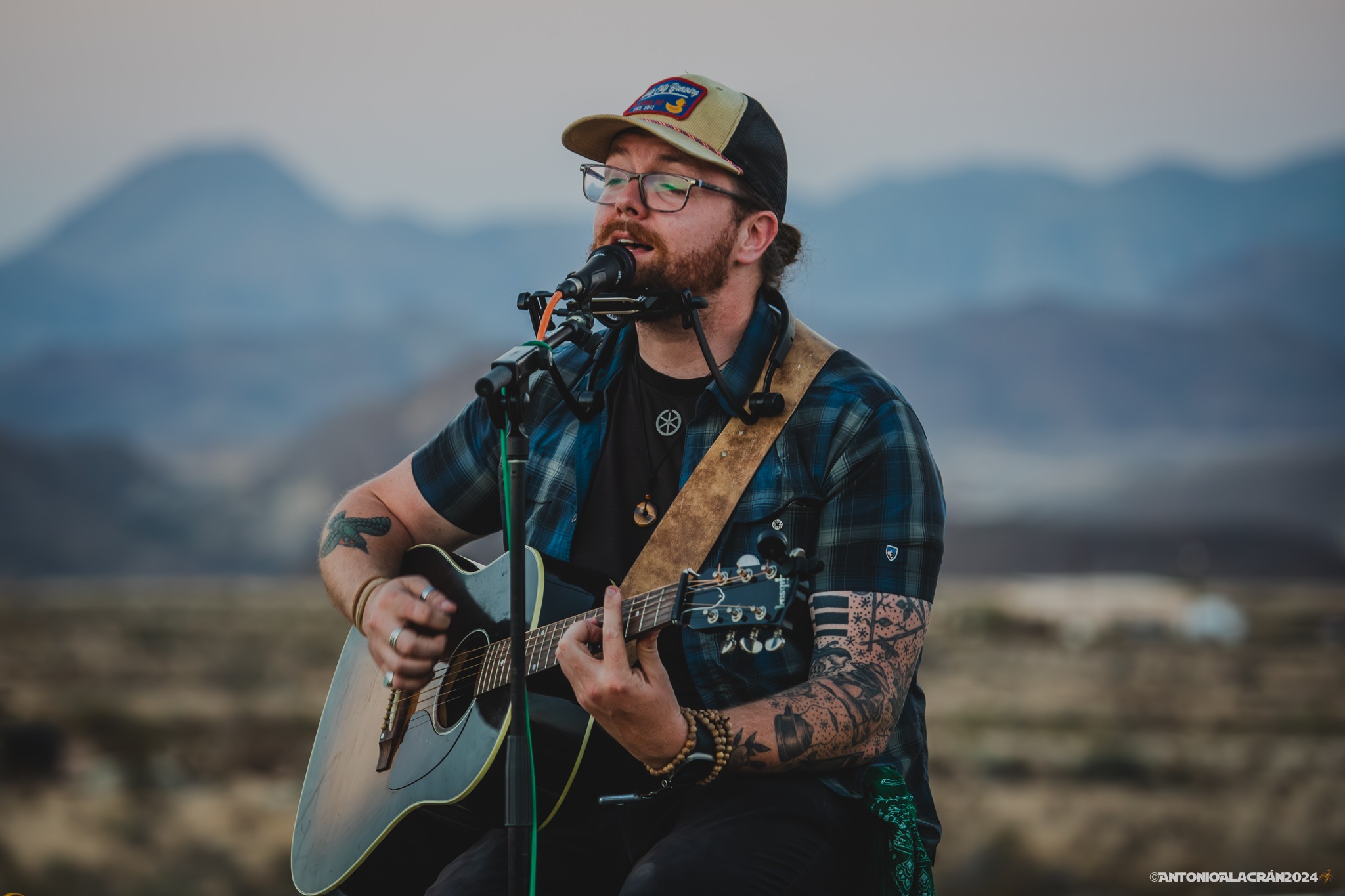Person with glasses and tattoos playing guitar and singing outdoors with mountains in background.