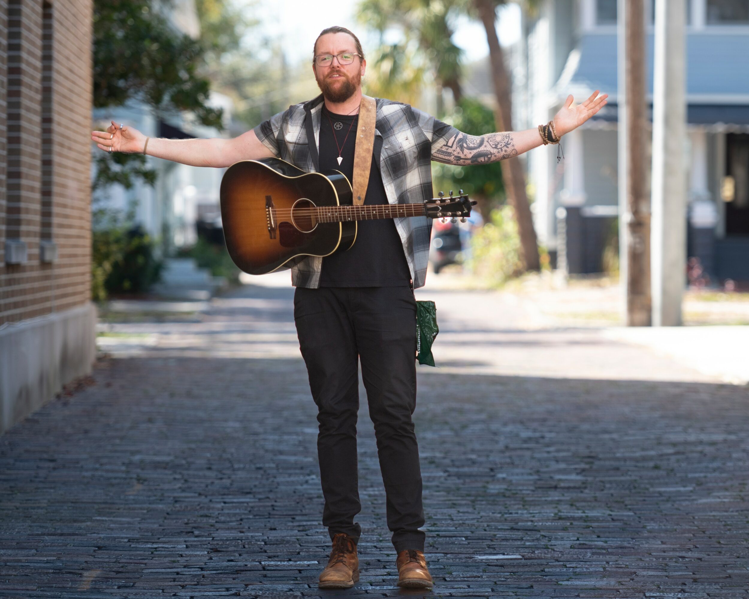 Man with a guitar standing outdoors with arms outstretched, smiling, in an urban setting.