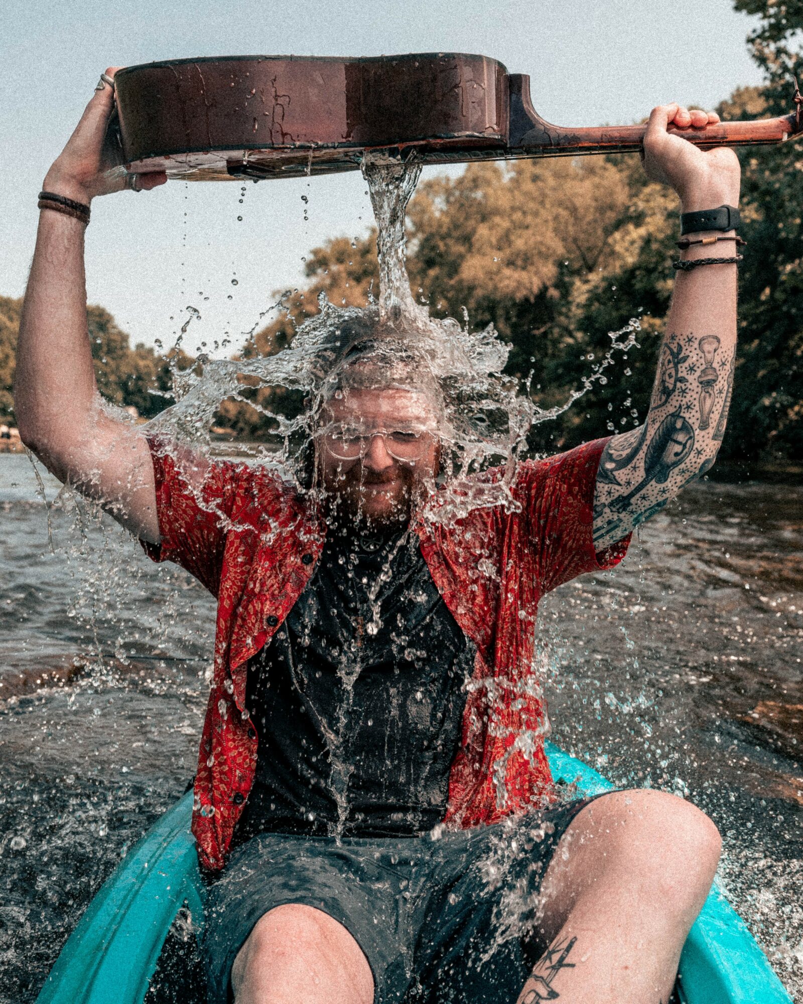 Person with tattoos and glasses holding a large container pouring water over their head outdoors.