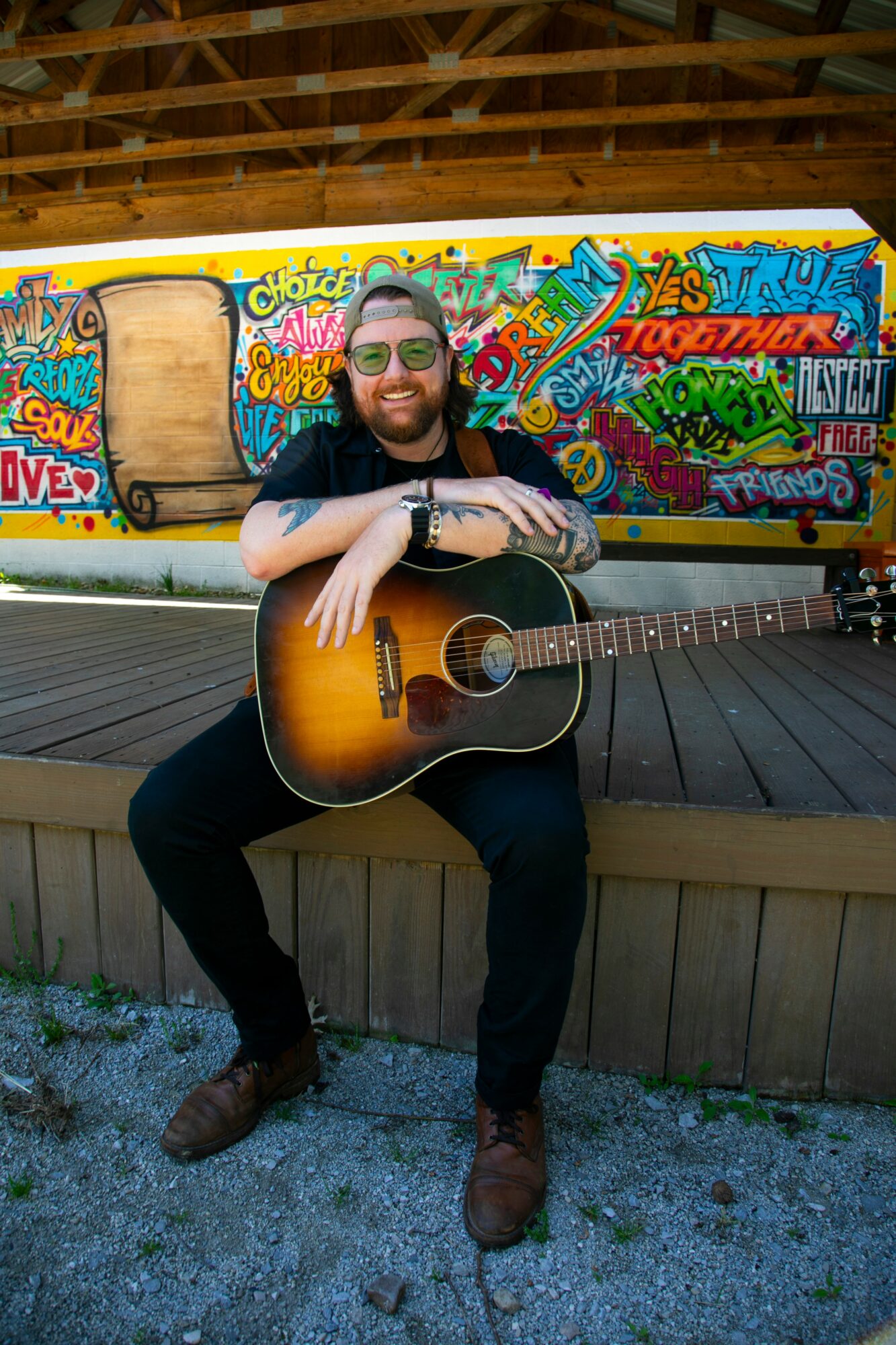 Person with sunglasses sitting on a wooden platform holding an acoustic guitar, graffiti-covered wall behind them.