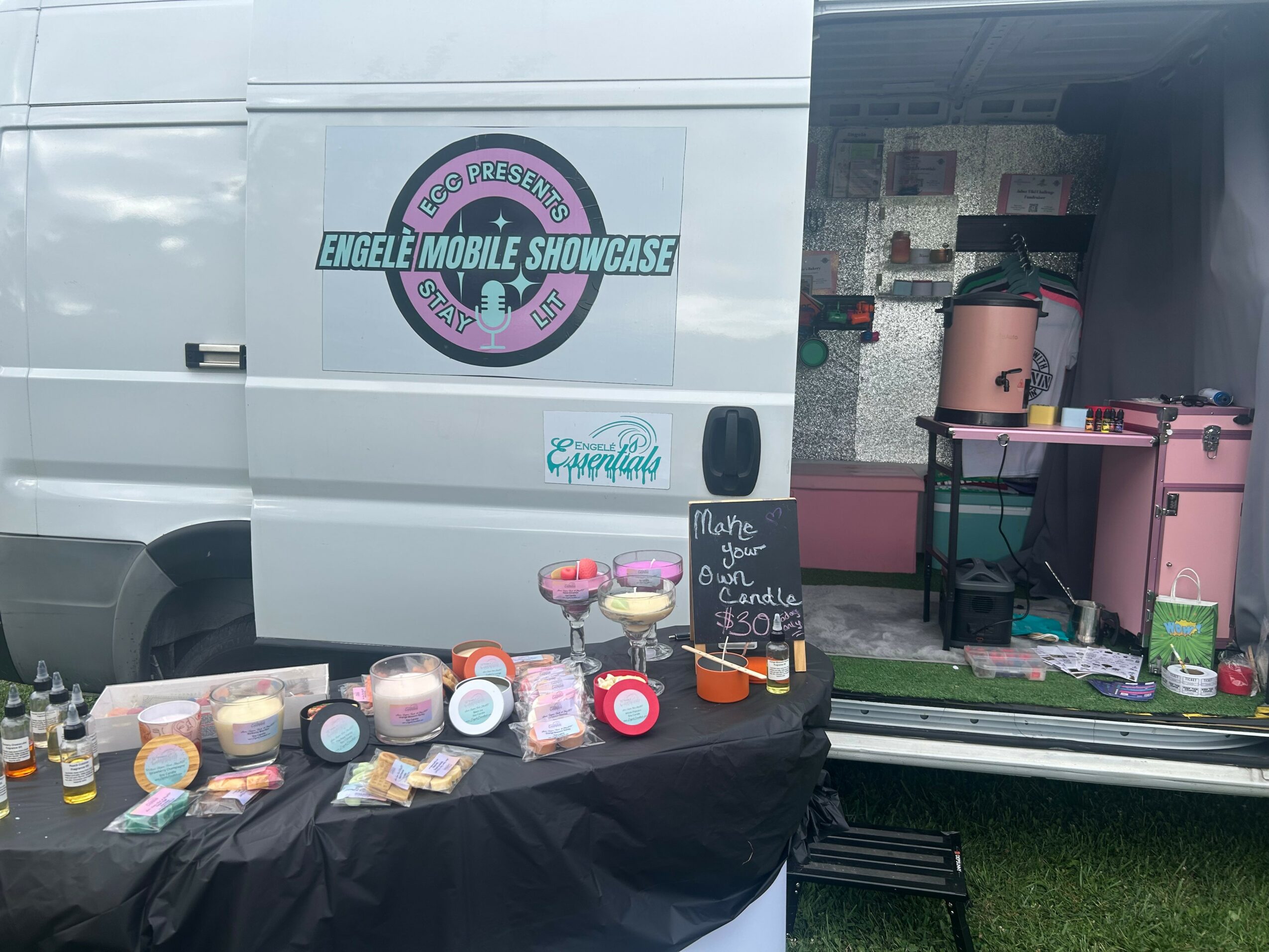 Display table with colorful desserts and a chalkboard sign, set up outside a mobile showcase van with pink equipment.