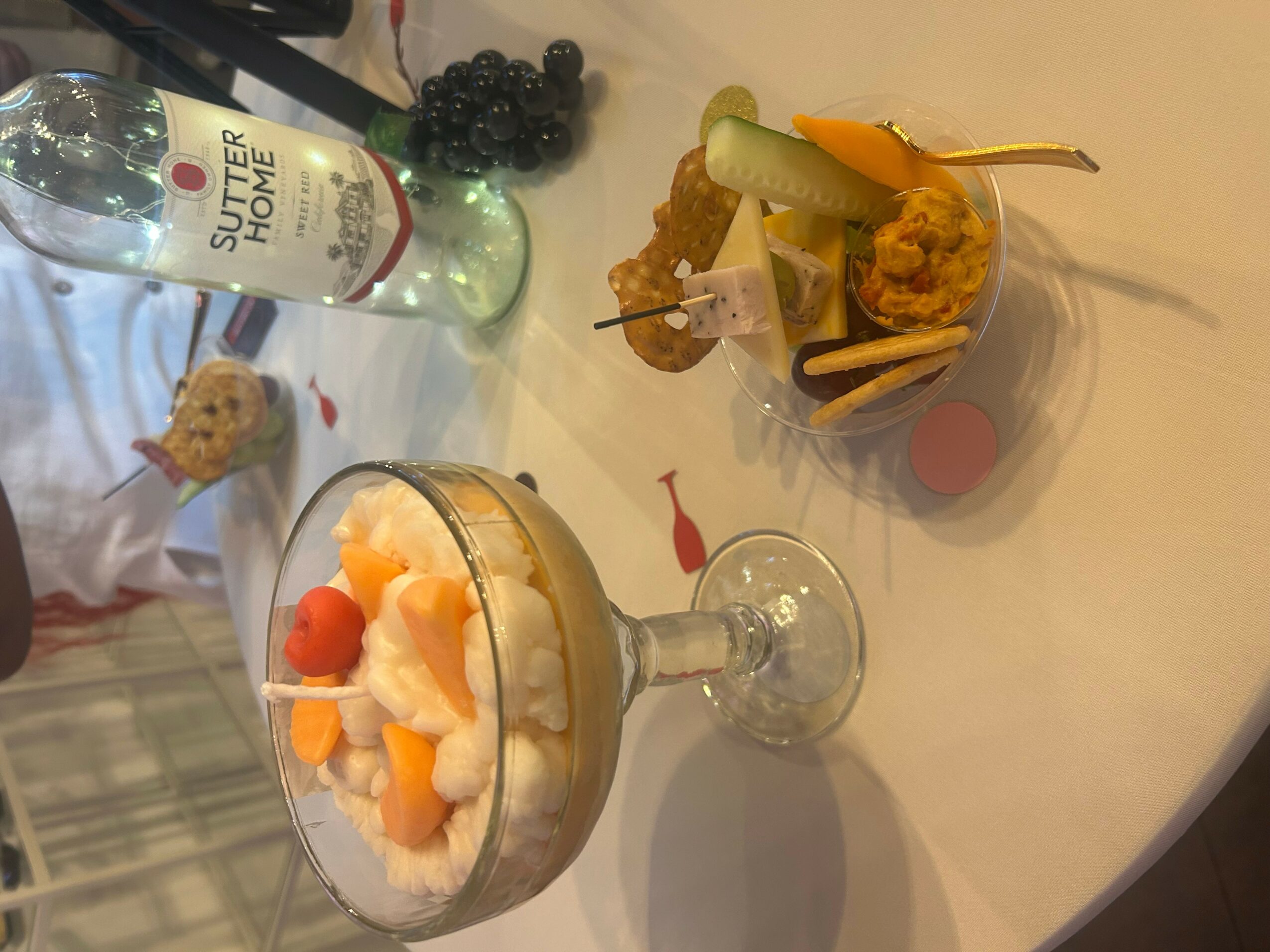 Dessert in a glass bowl with fruit, a bottle of water, and a small bowl with fried food and pickles on a table.
