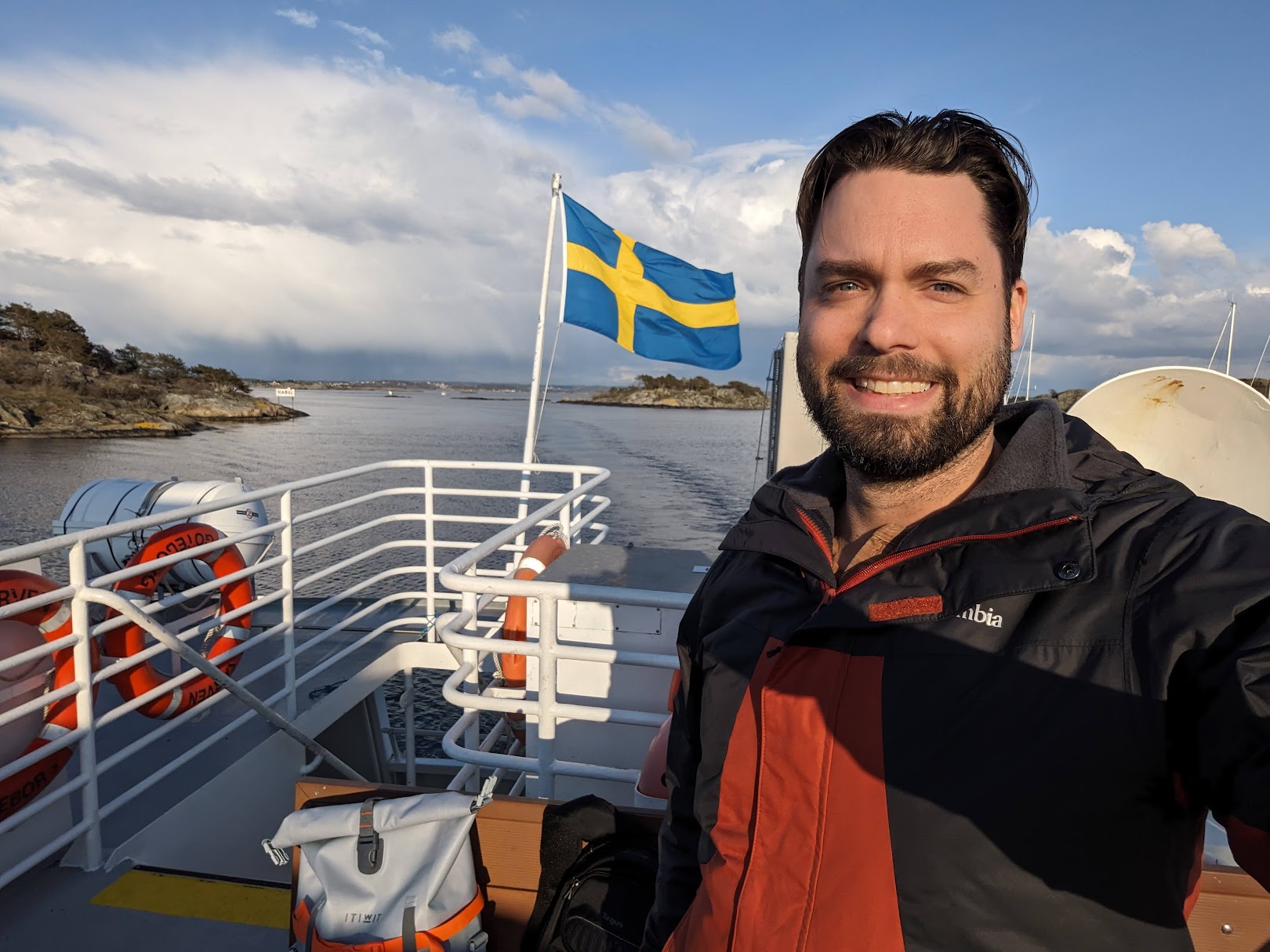 Man with dark hair and beard smiling on boat with Swedish flag, water, and sky in background.