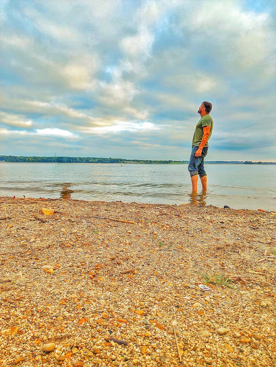 Person standing in water at the beach, looking at the sky with clouds, sandy shore in foreground.